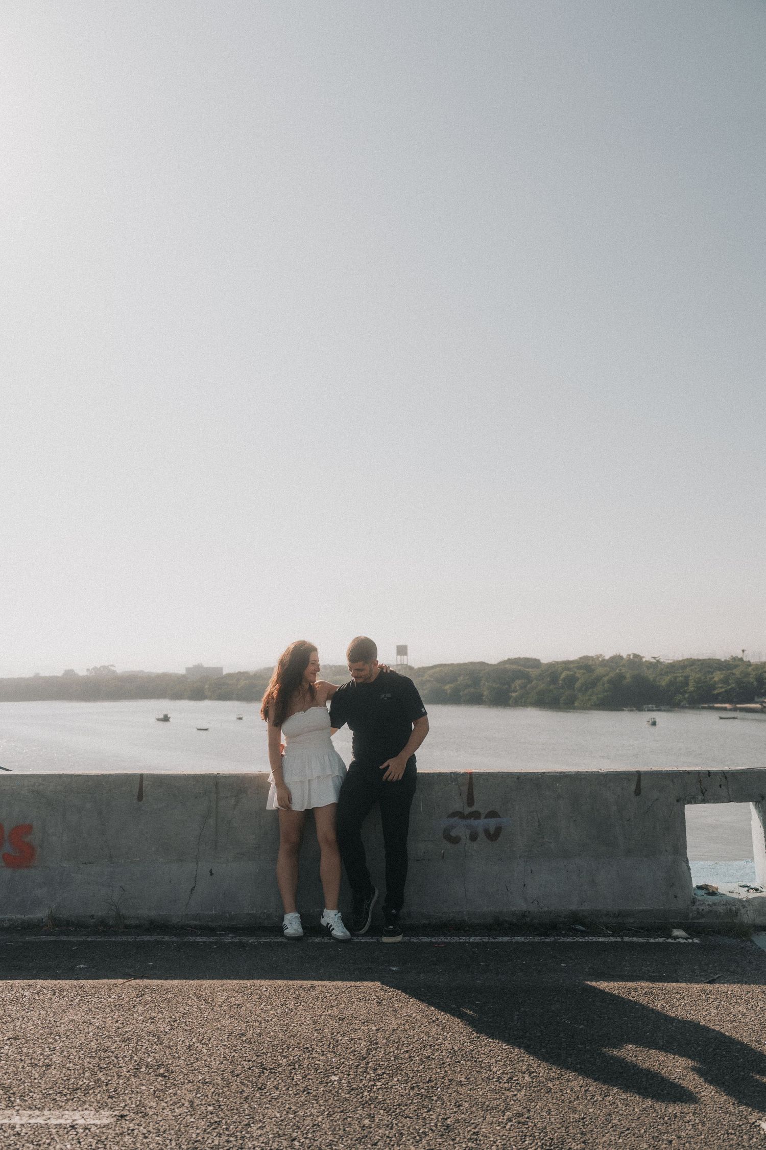 Two people lean against a stone wall overlooking a distant harbor on a sunny summer day.