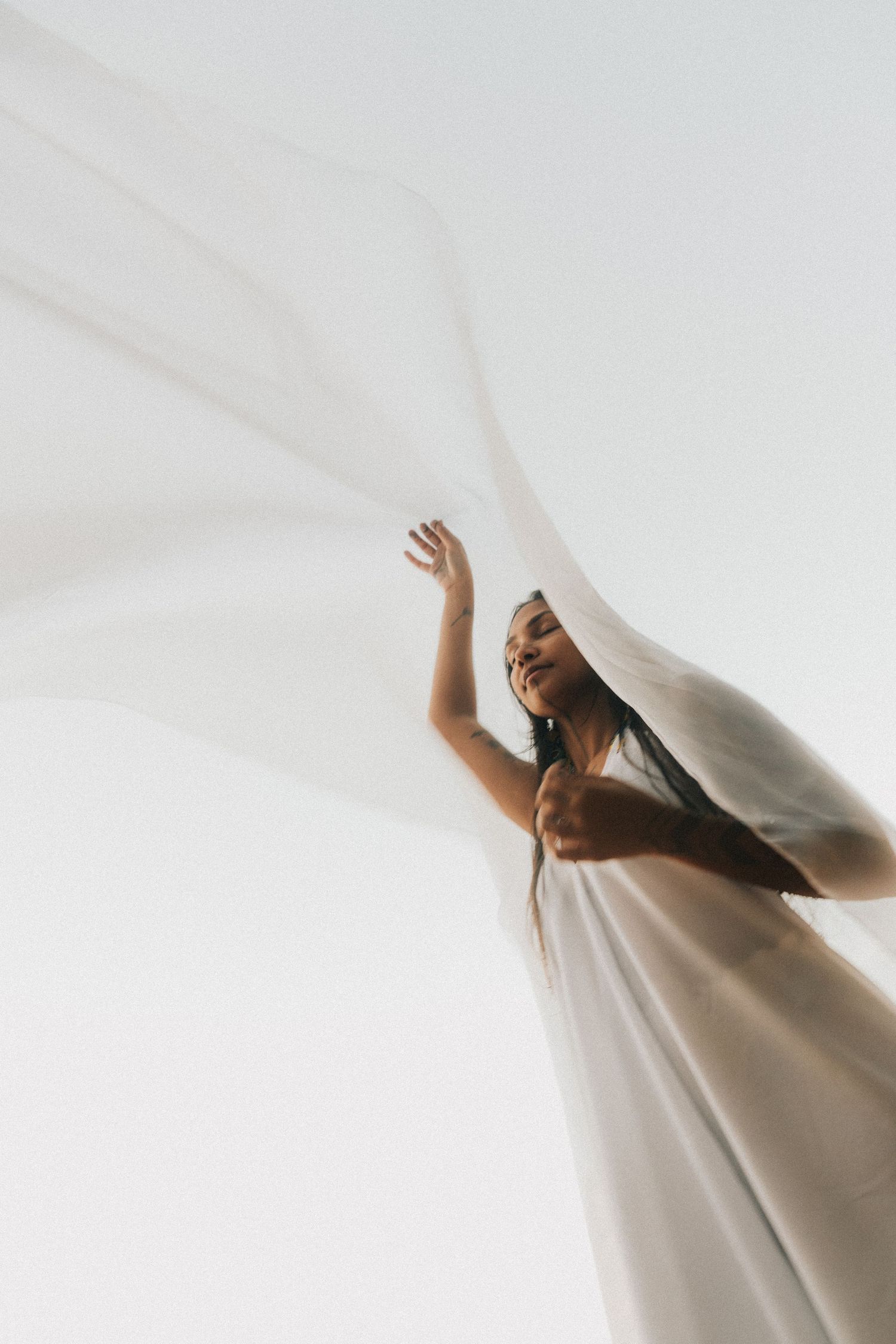 Figure in flowing white fabric reaches upward against bright background creating ethereal artistic effect.