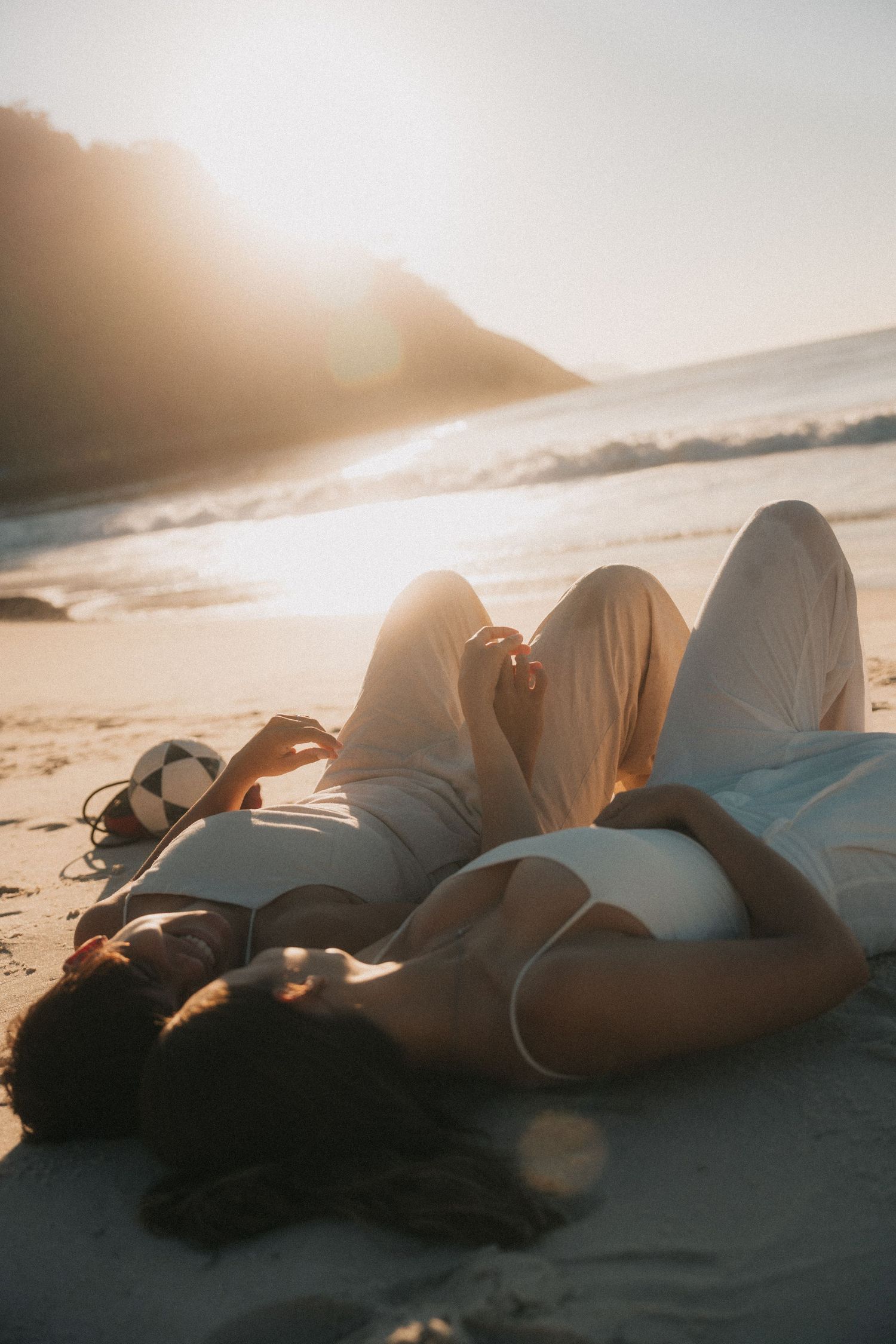 Silhouetted figures lying comfortably on sandy beach during golden sunset by the ocean.