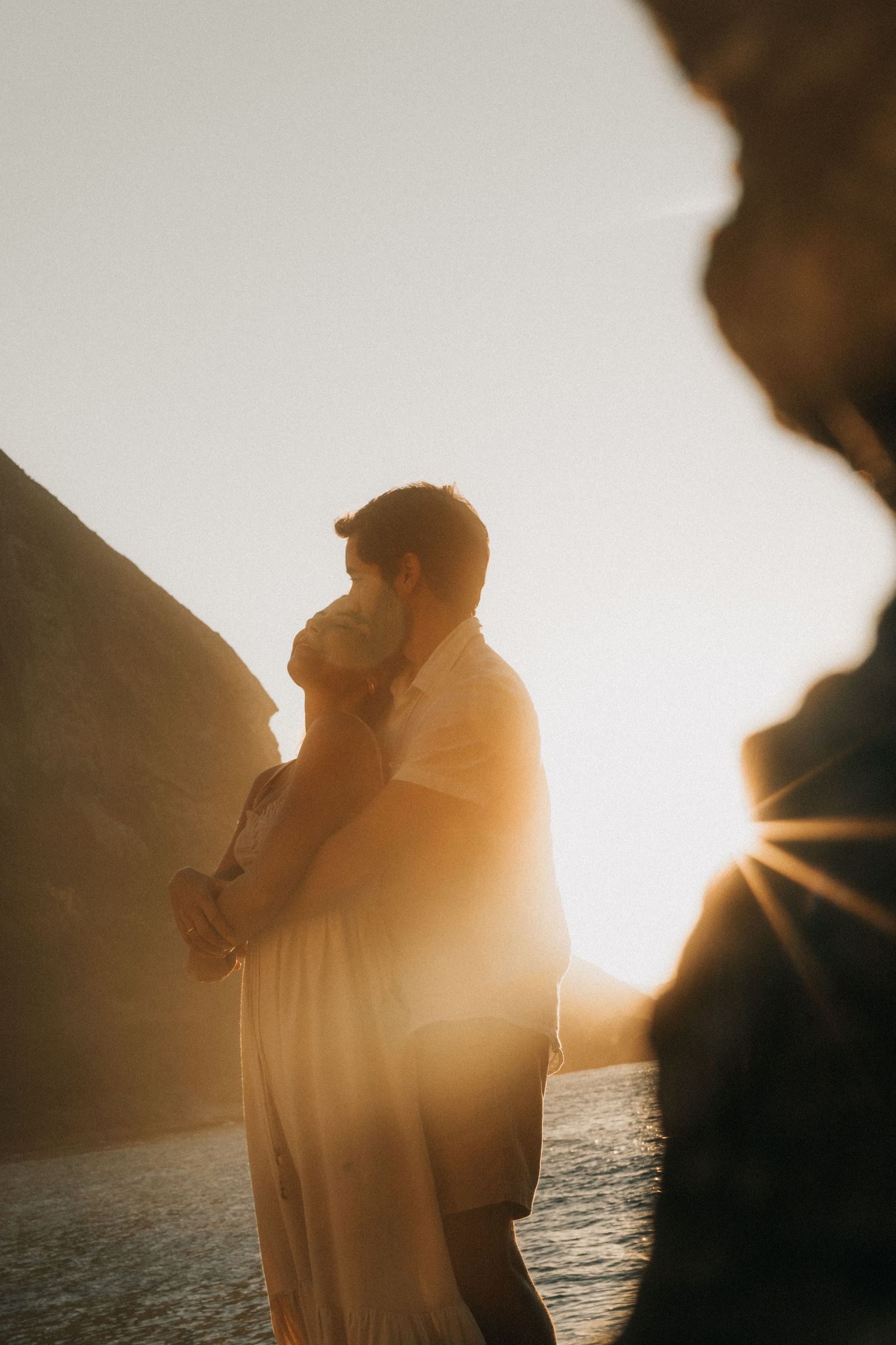 Silhouetted couple embraces against golden sunset over ocean cliffs, creating romantic backlit scene.