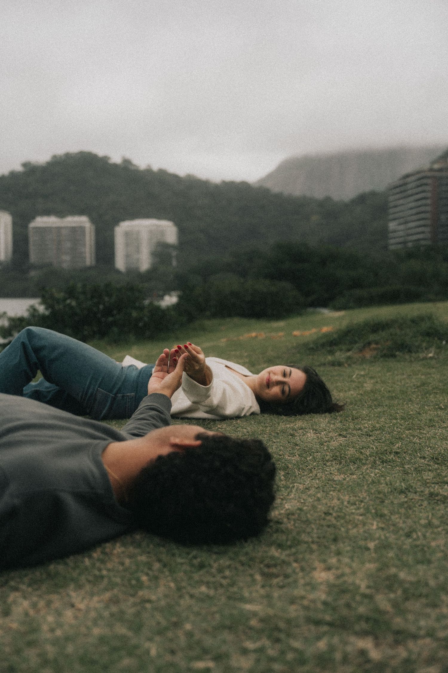 Two people lying down on a green grassy hilltop with city buildings visible in the foggy mountain background.
