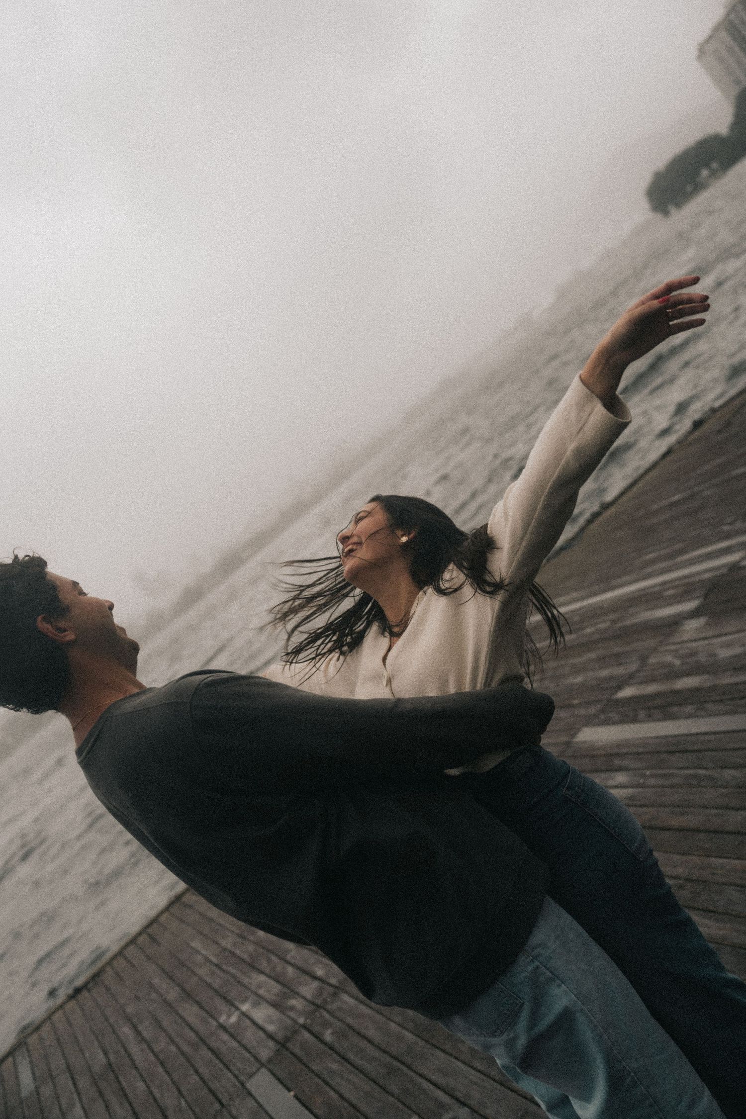 Two dancers perform an expressive contemporary dance move on a wooden walkway in moody overcast weather.