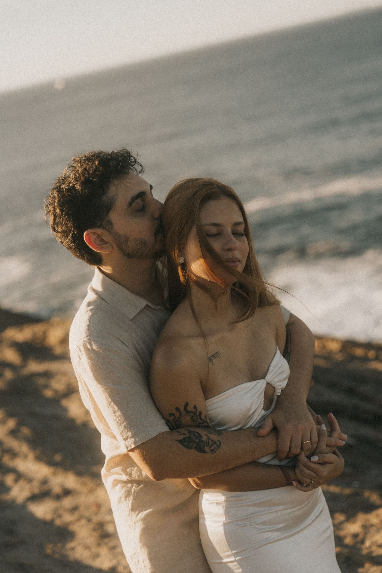 A couple shares romantic moments on a beach at sunset with waves crashing in the background.