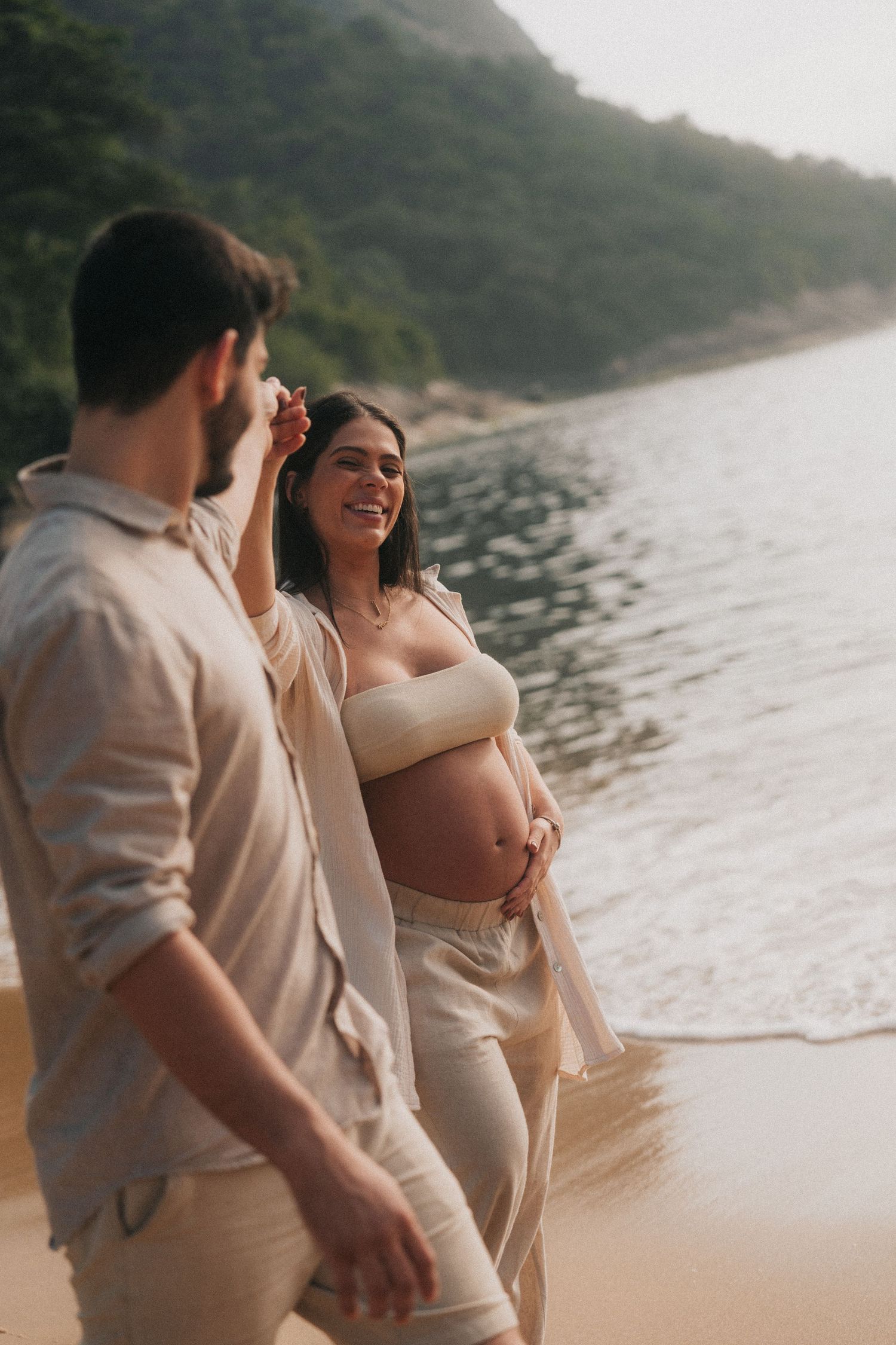 Expectant couple sharing a joyful moment on a beach with mountains in the background.