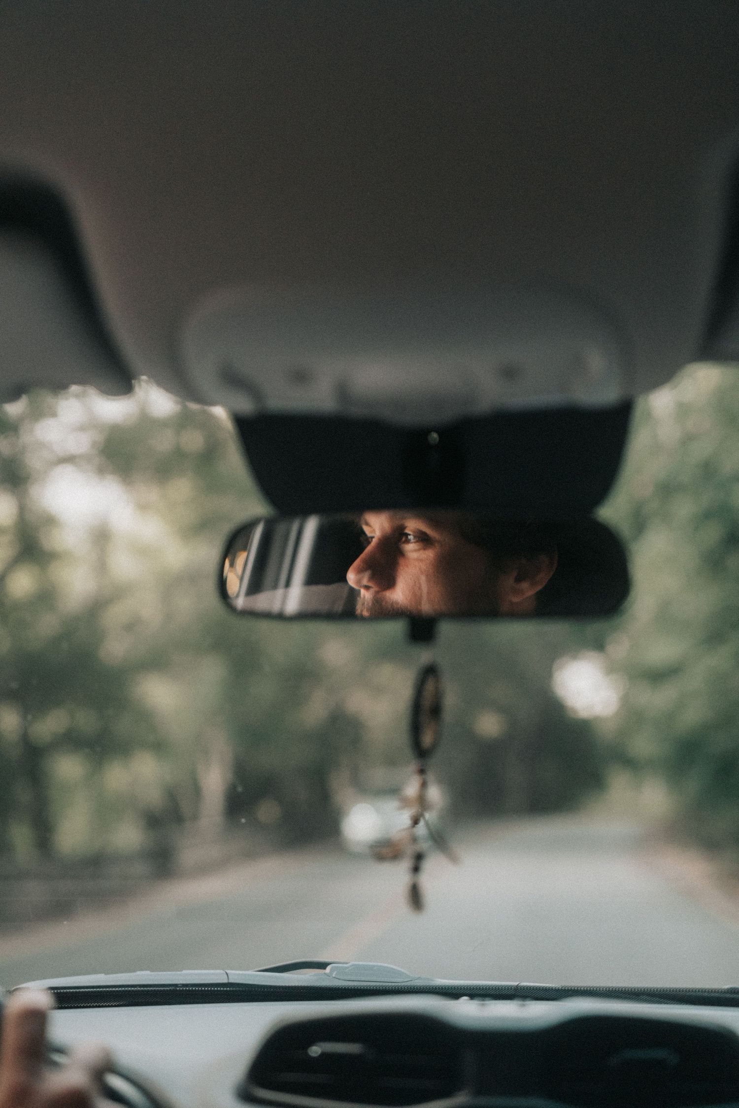 Car interior view through windshield showing road ahead and rear view mirror against blurred natural background.