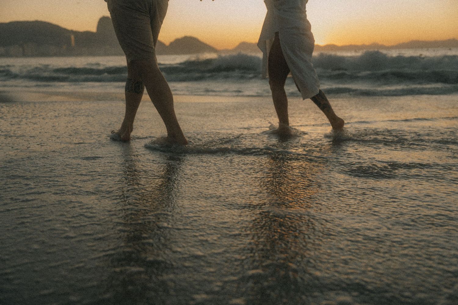 Silhouetted legs and feet walk along a sandy beach at sunset with ocean waves rolling in the background.
