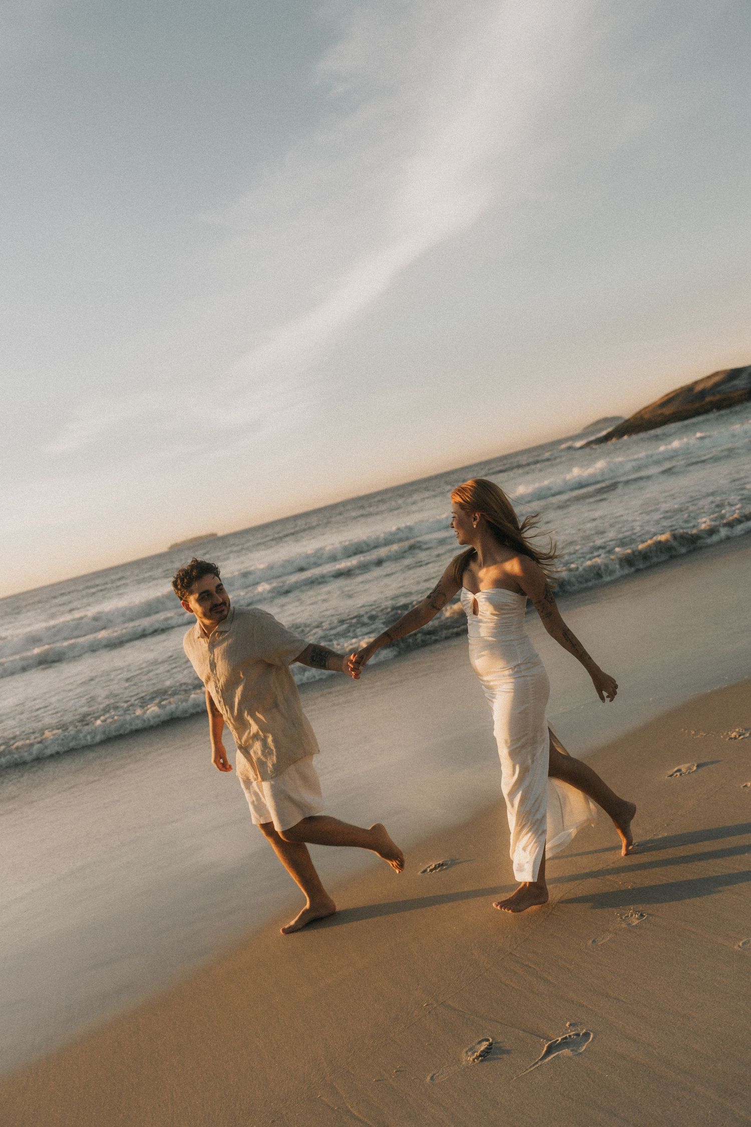 Two people in white clothing playfully dance together on a sandy beach at sunset while ocean waves roll in.