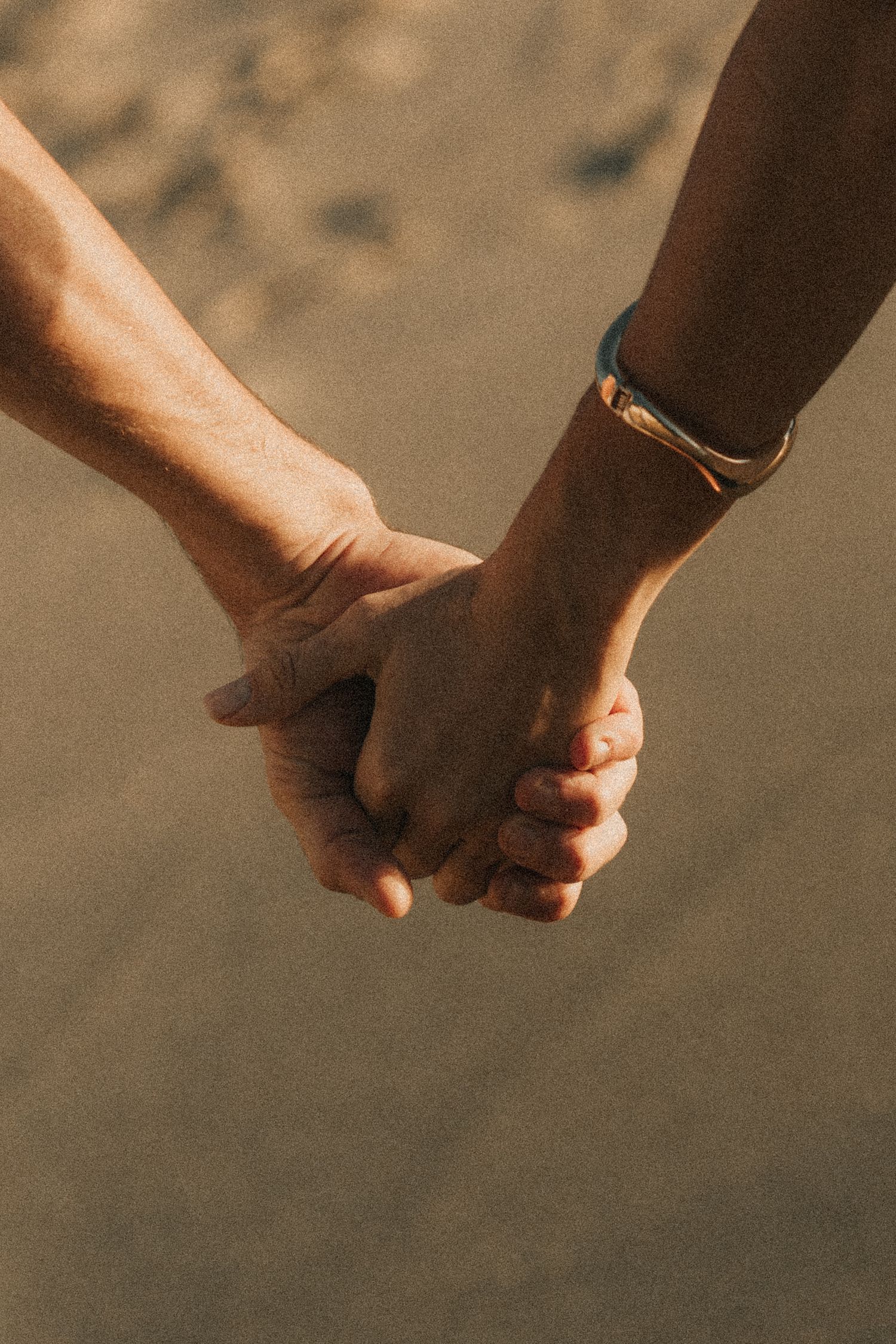 Two different-toned hands gently hold each other in closeup against a blurred neutral background.