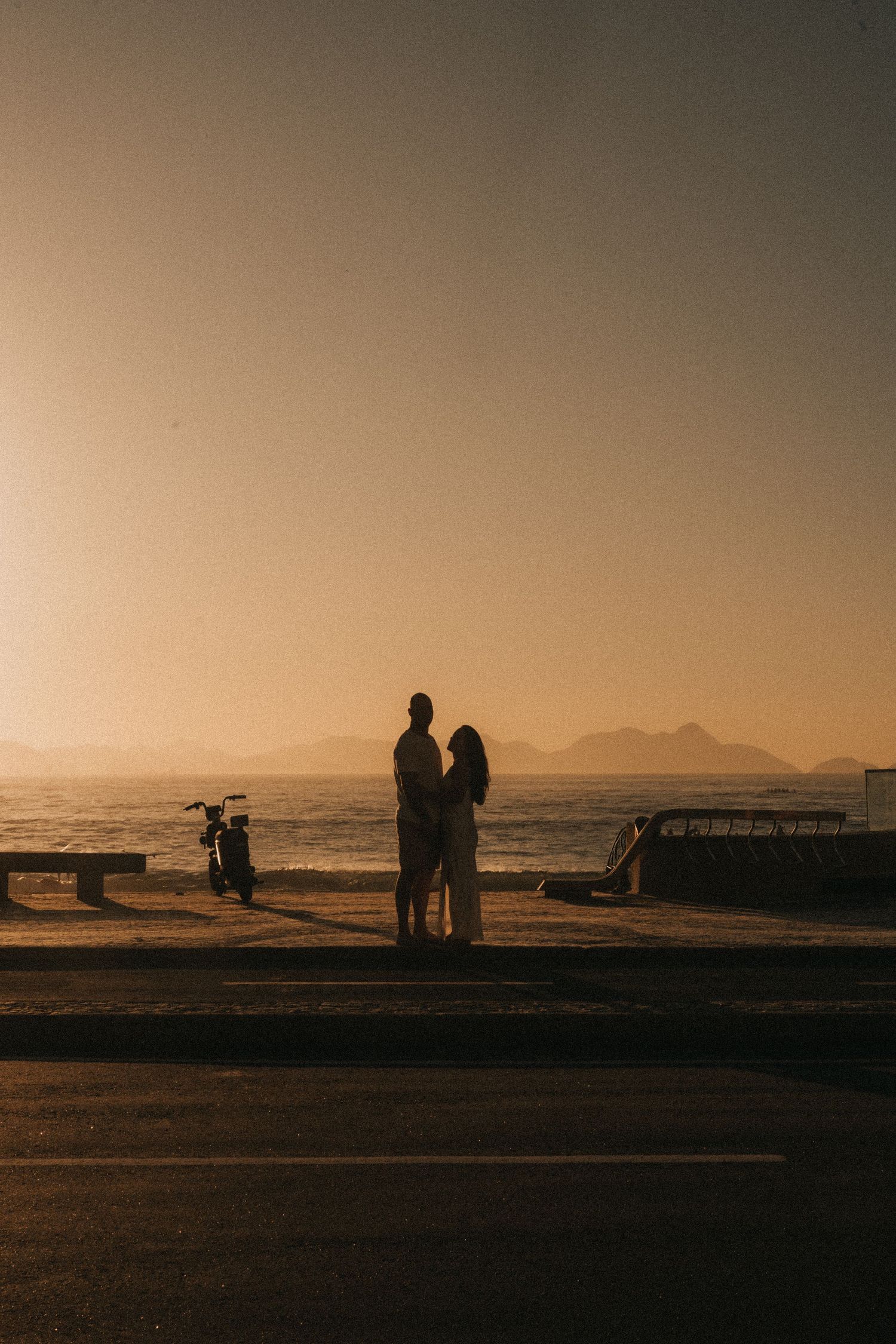 Silhouettes of people walking on a beach walkway at sunset with mountains in the distance.