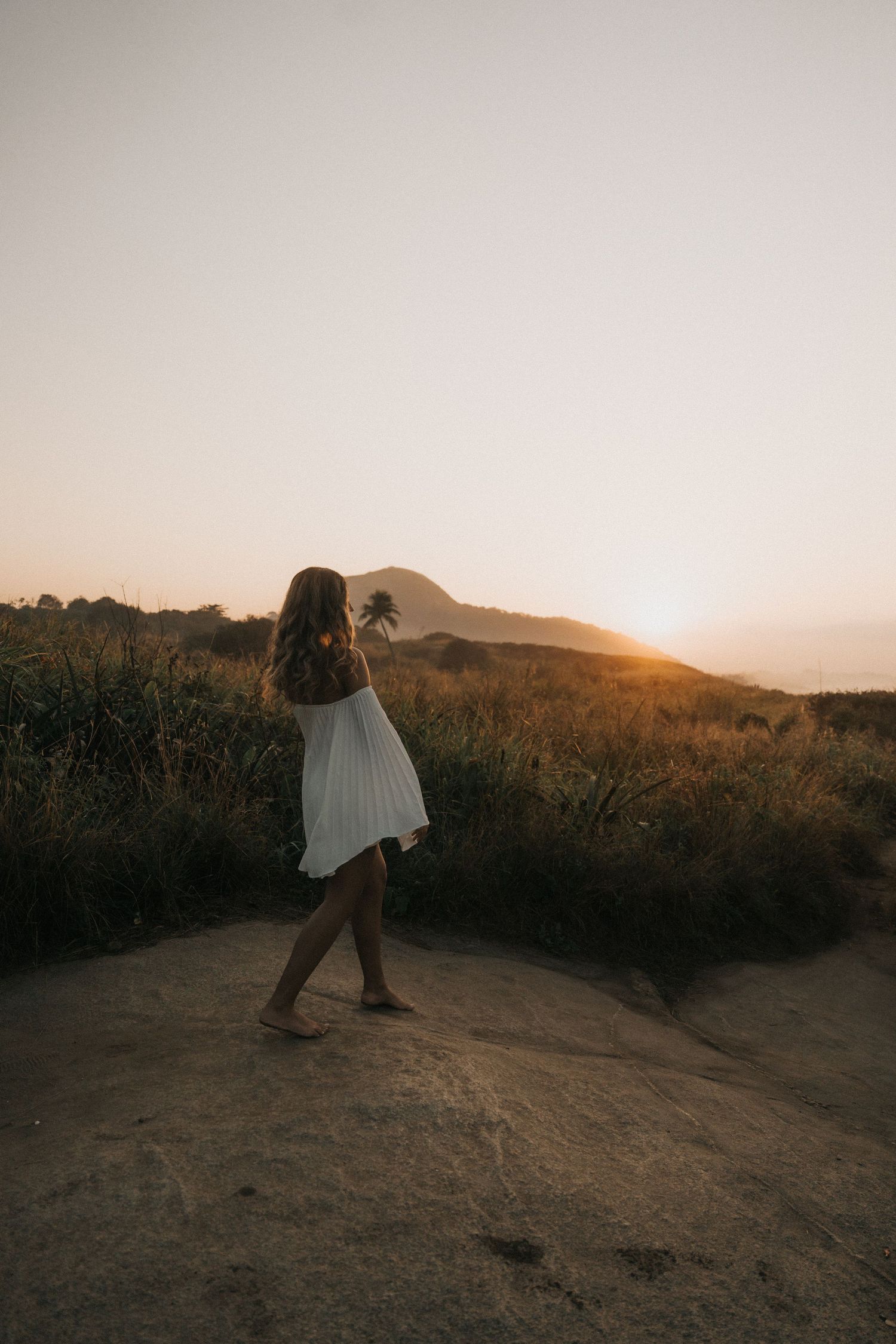 A person wearing a white dress stands on a mountain trail against a golden sunset silhouetting distant hills.