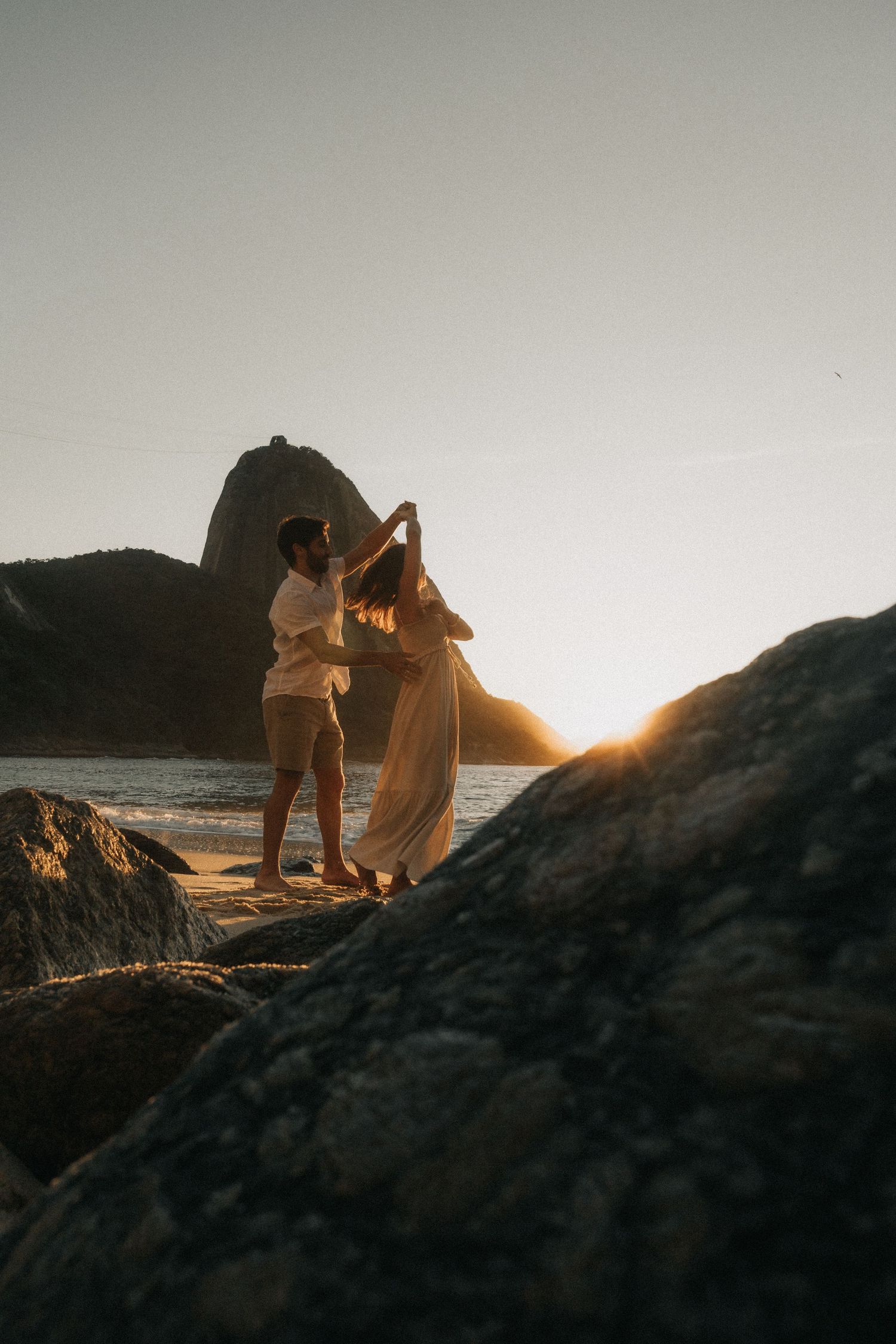 Two figures embrace on rocky coastal cliffs against a dramatic golden sunset over the ocean.