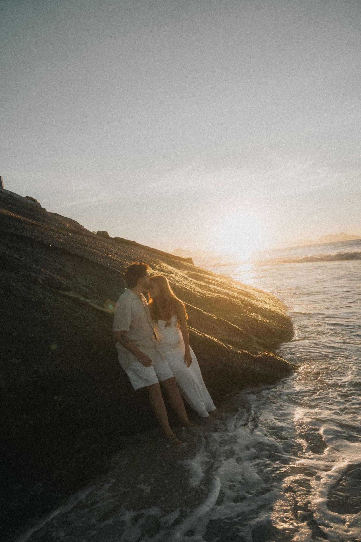 A couple sits on coastal rocks at sunset as ocean waves crash around them, creating a romantic silhouetted scene.