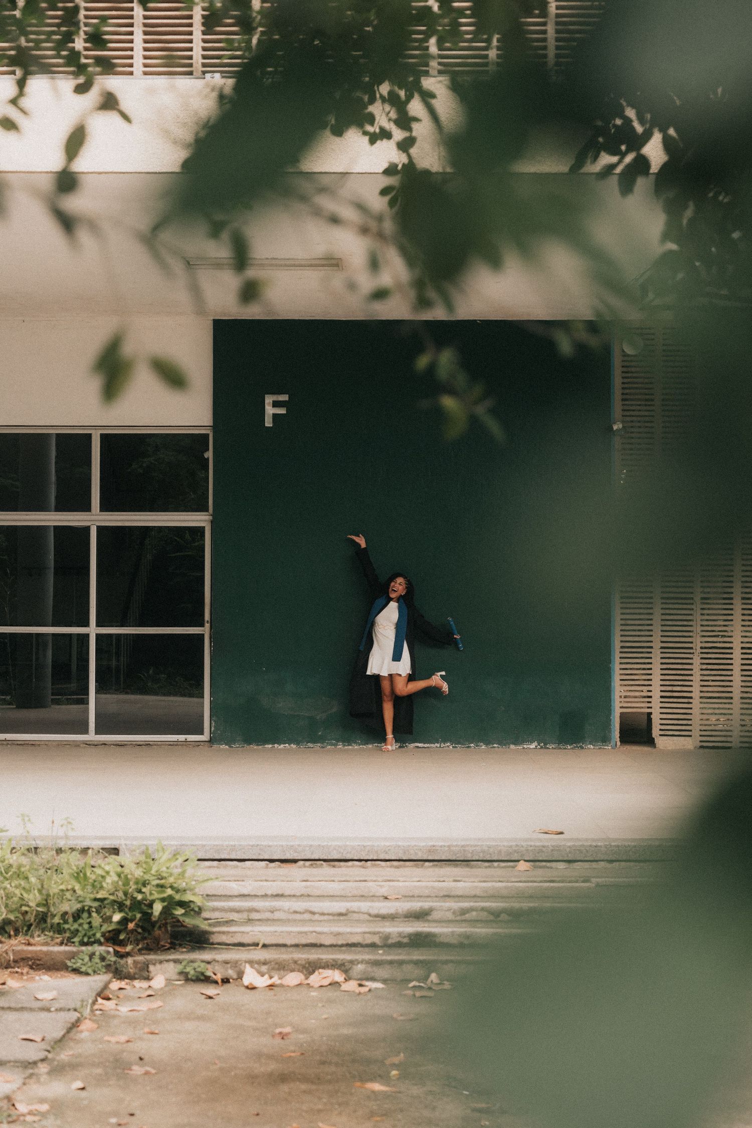 Through foliage, a distant figure stands against a light green wall with windows in an urban setting.
