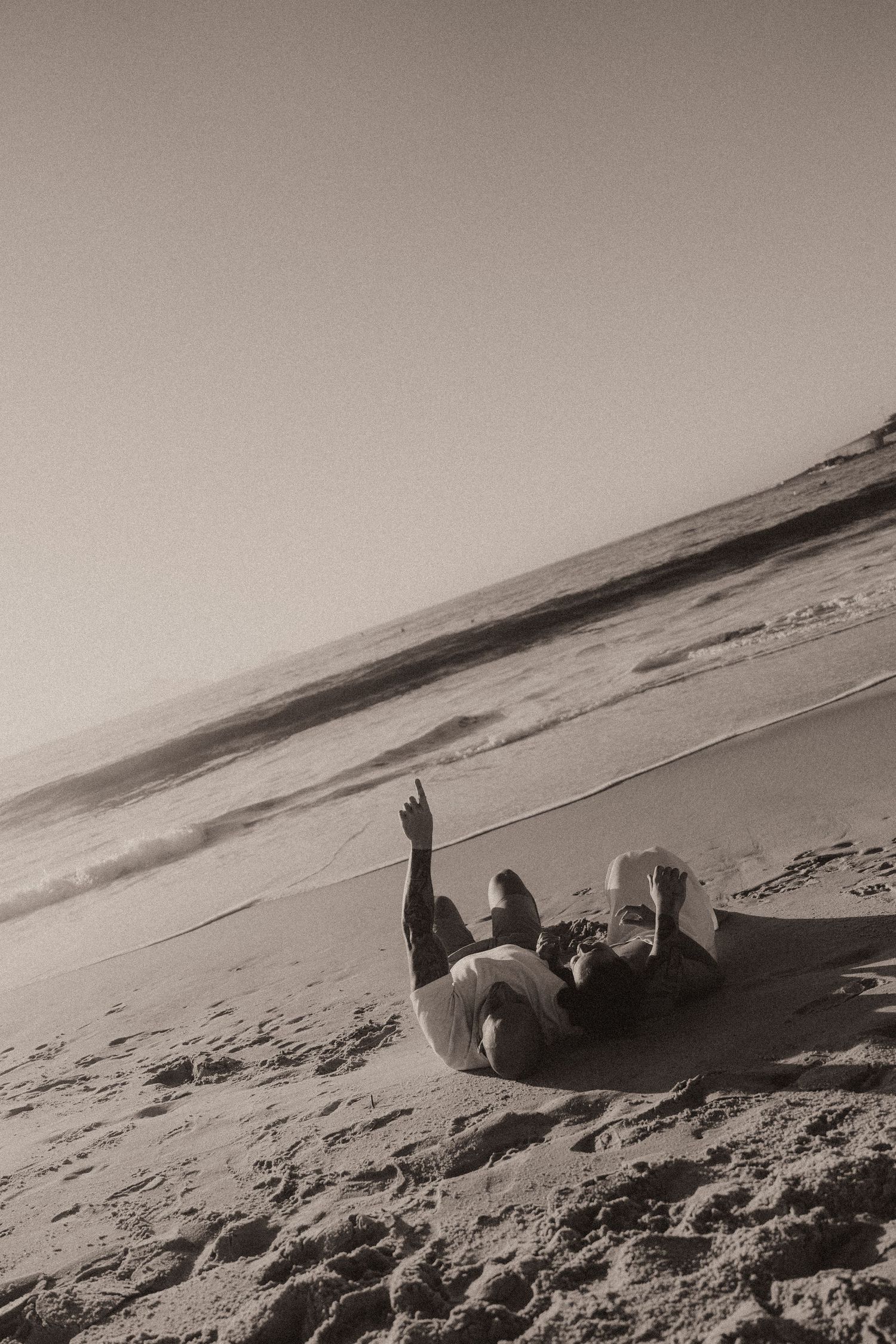 A figure lies in black and white on a sandy beach slope with an outstretched arm against a serene coastal backdrop.