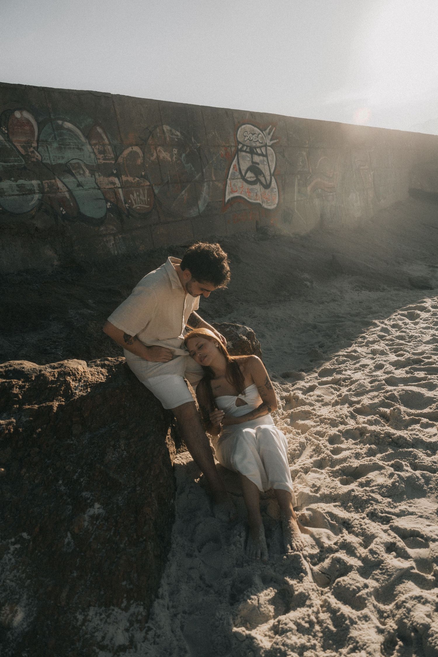 A loving couple embraces on a sandy beach with graffiti-covered rocks at sunset creating a romantic silhouette.