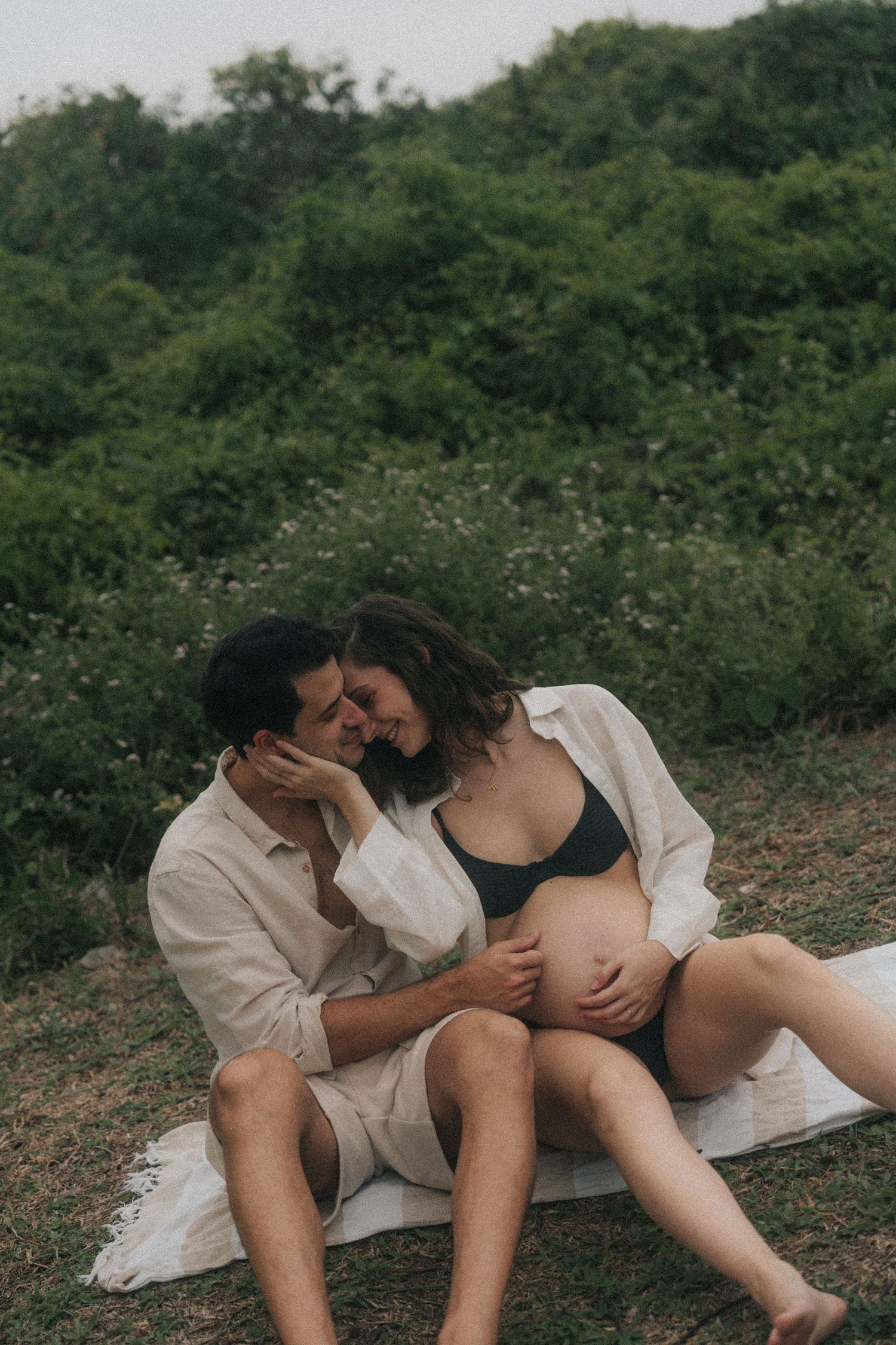 An intimate maternity photo shoot outdoors with a couple sitting on a white blanket.