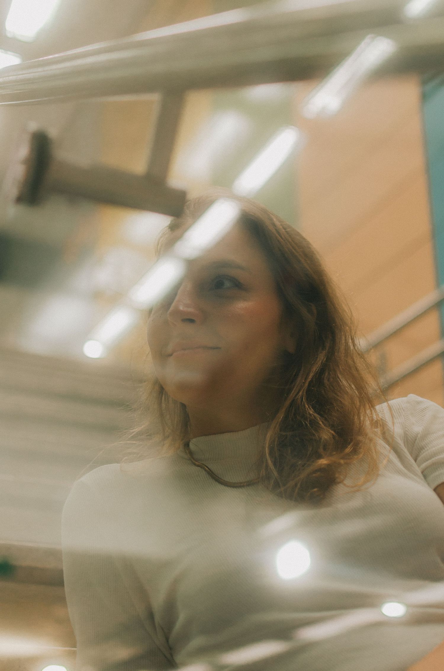 A person in a white turtleneck sweater looking upward with a soft smile in warm lighting with overhead fluorescent lights.