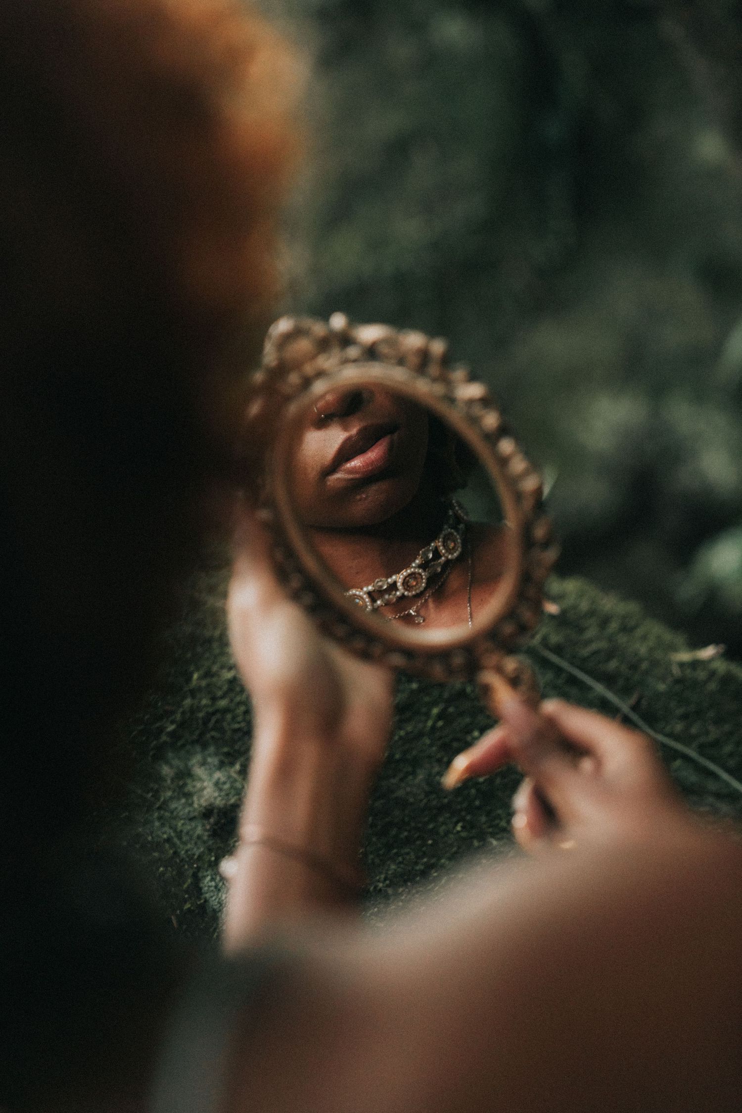 A vintage circular mirror held in nature reflects lips wearing a chain necklace in an artistic portrait composition.