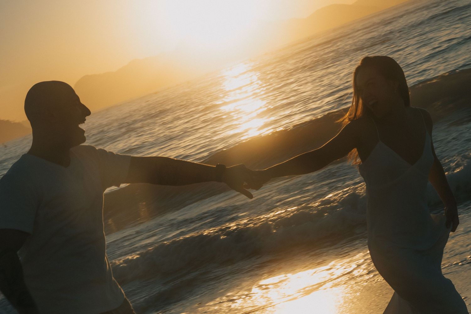 Silhouetted figures stand along a beach shoreline at golden sunset as waves crash against the shore.