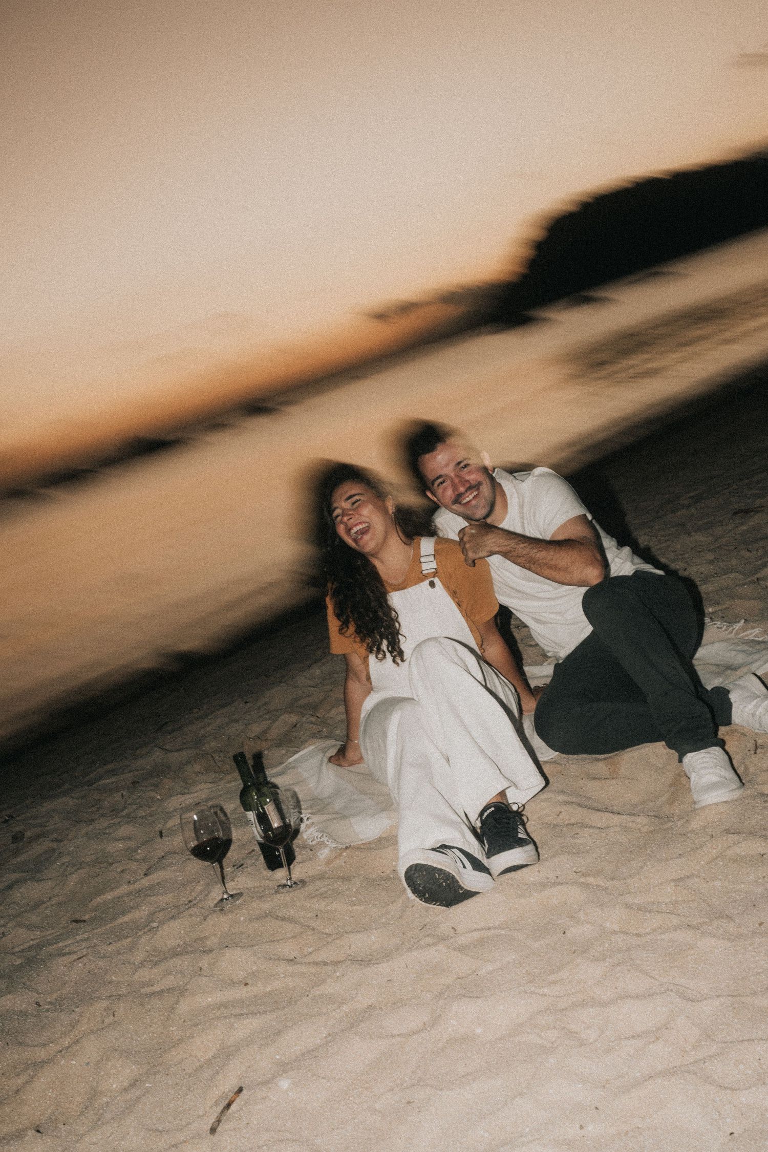 Two people in formal attire playfully roll around on the sandy beach during a sunset photoshoot.