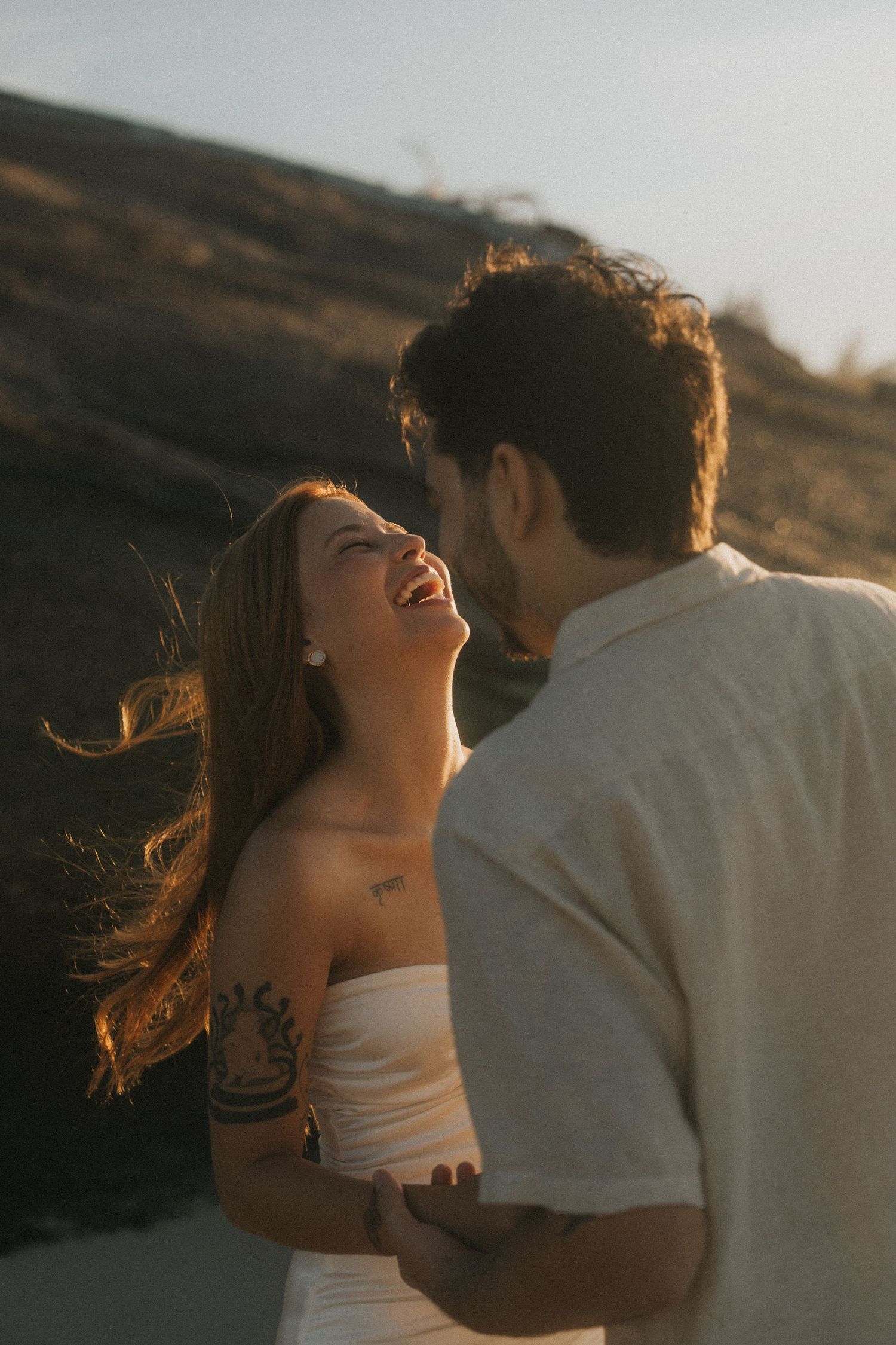 A couple shares an intimate moment at sunset with warm golden light illuminating their silhouettes against a hillside.