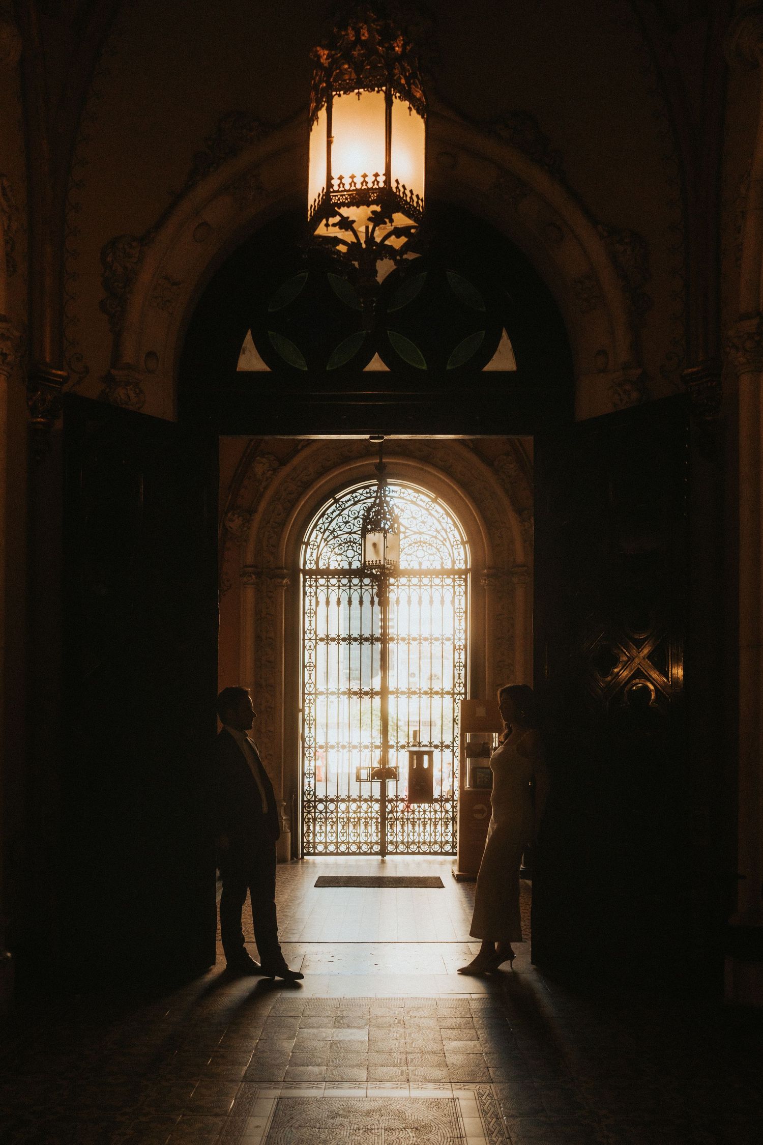 Silhouetted figures stand in a dramatic arched hallway with backlit windows creating moody architectural lighting.