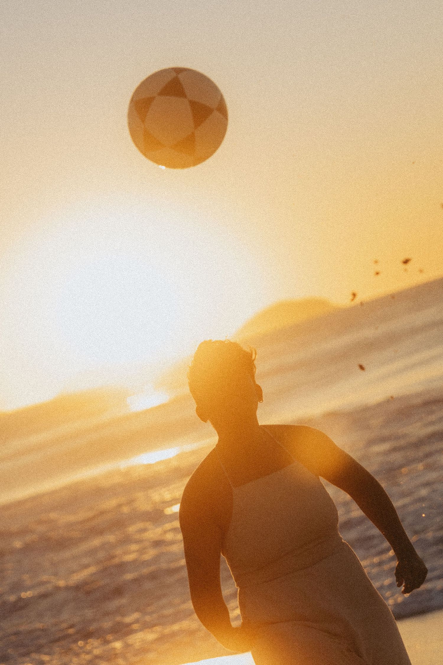 A silhouetted figure stands on a beach at sunset with an orange glowing sun and ocean in the background.
