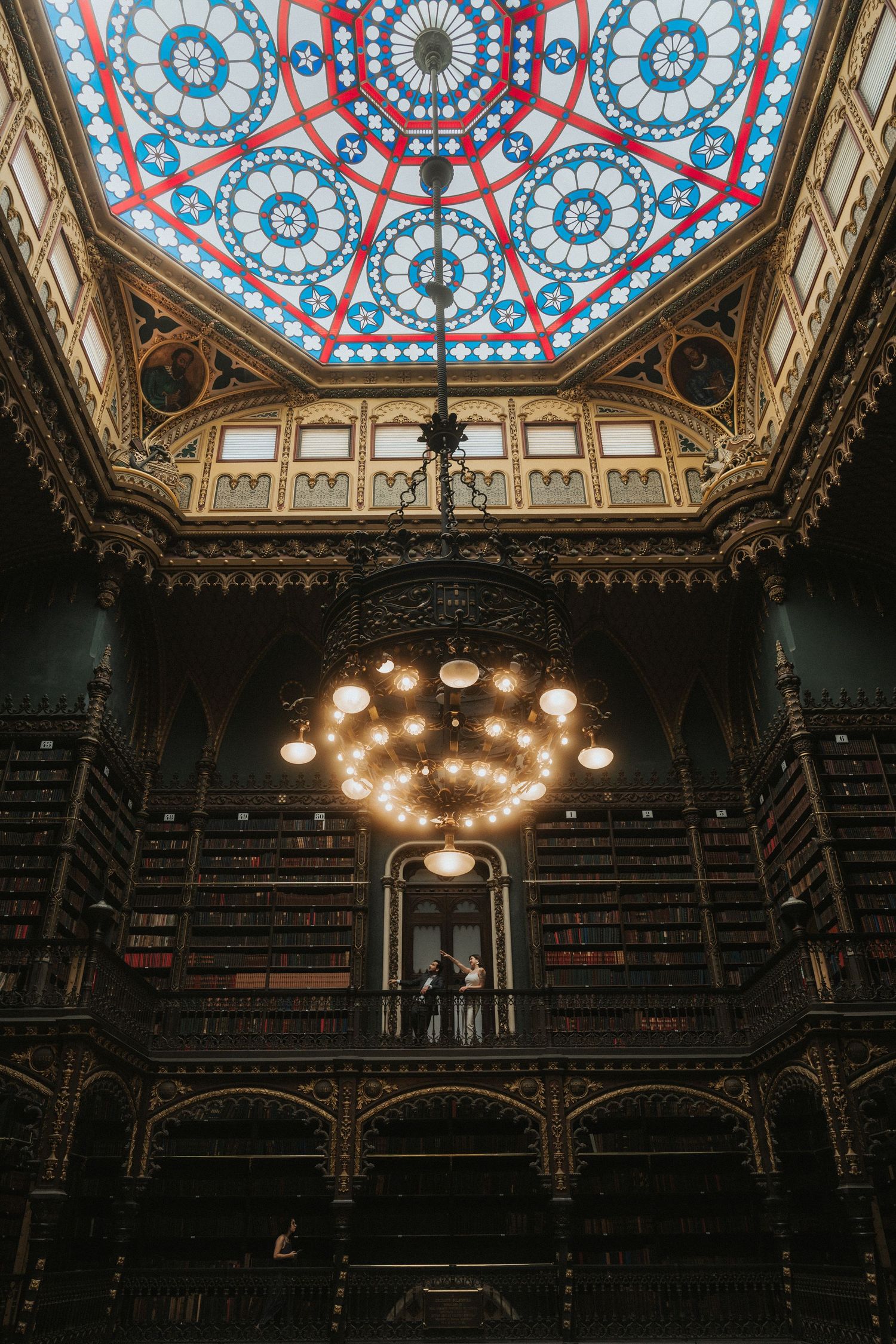 Ornate Victorian library interior with stained glass ceiling and grand chandelier illuminating dark wooden bookshelves.