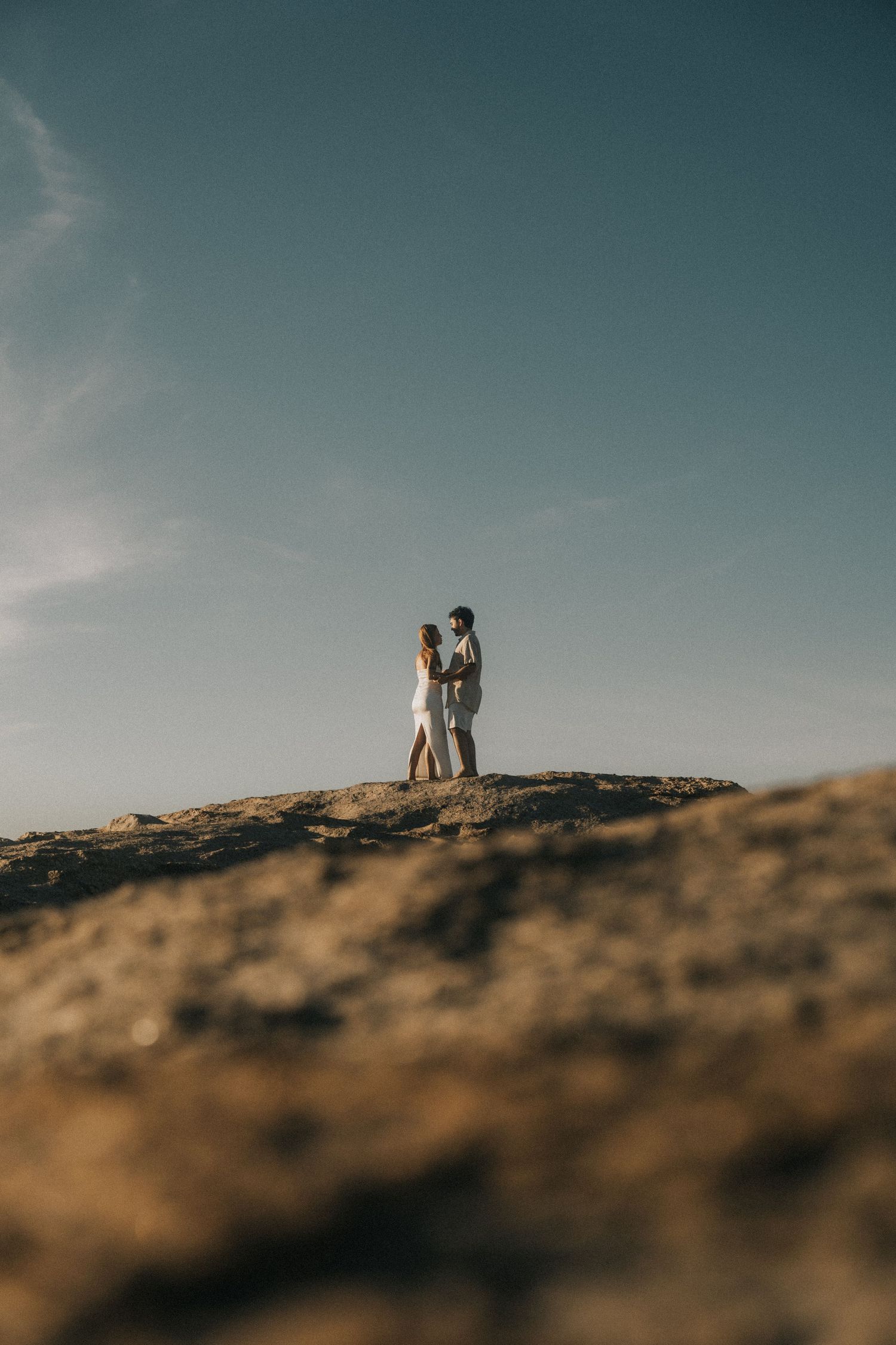 Two silhouettes stand together against a moody blue sky on a rocky hilltop at sunset.
