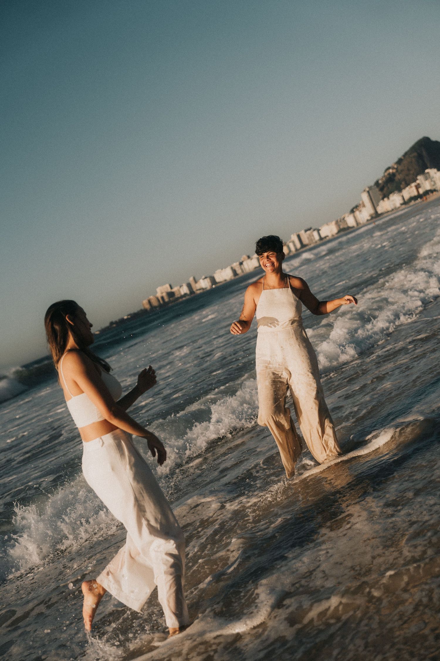 A romantic couple in white outfits plays and splashes together at the beach during sunset near coastal buildings.