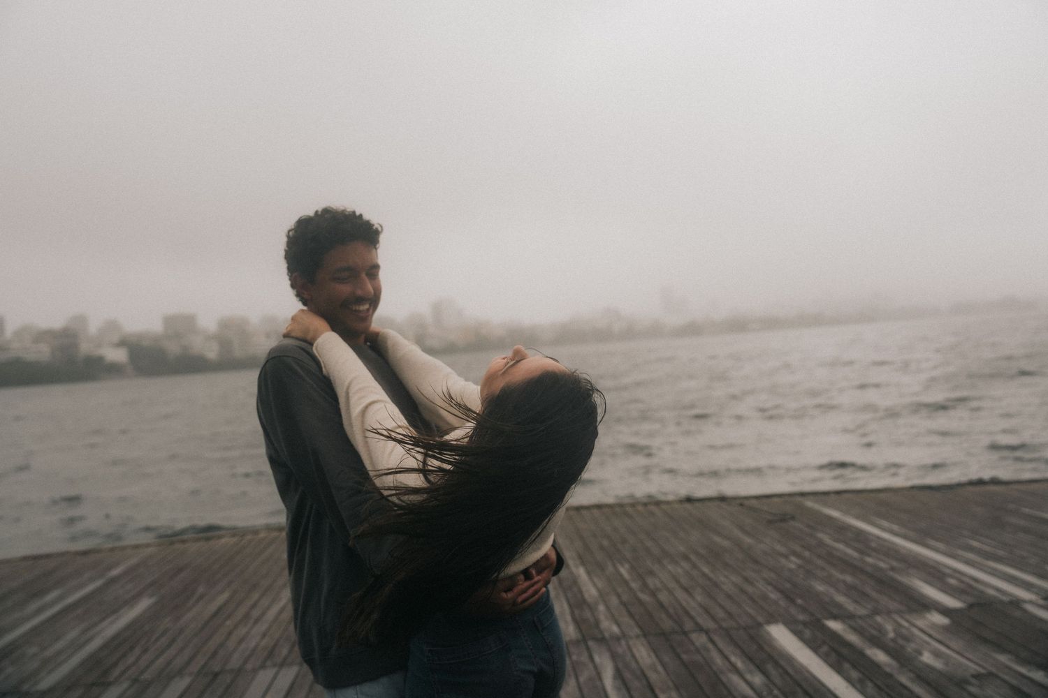 Couple sitting together on misty wooden pier by foggy lake during romantic moody morning.