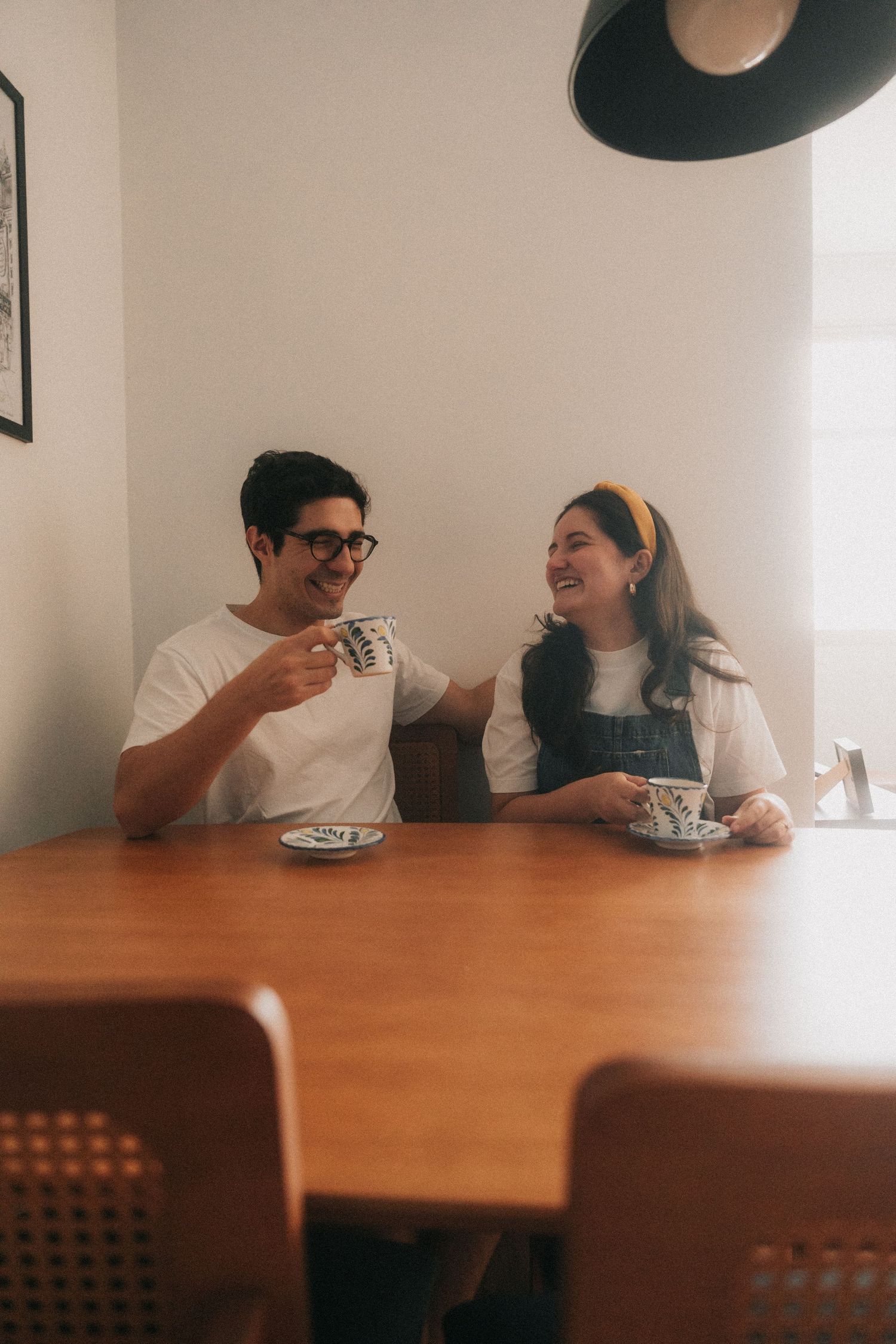 Two people share a joyful moment while having coffee and dessert at a wooden table in a minimalist dining space.