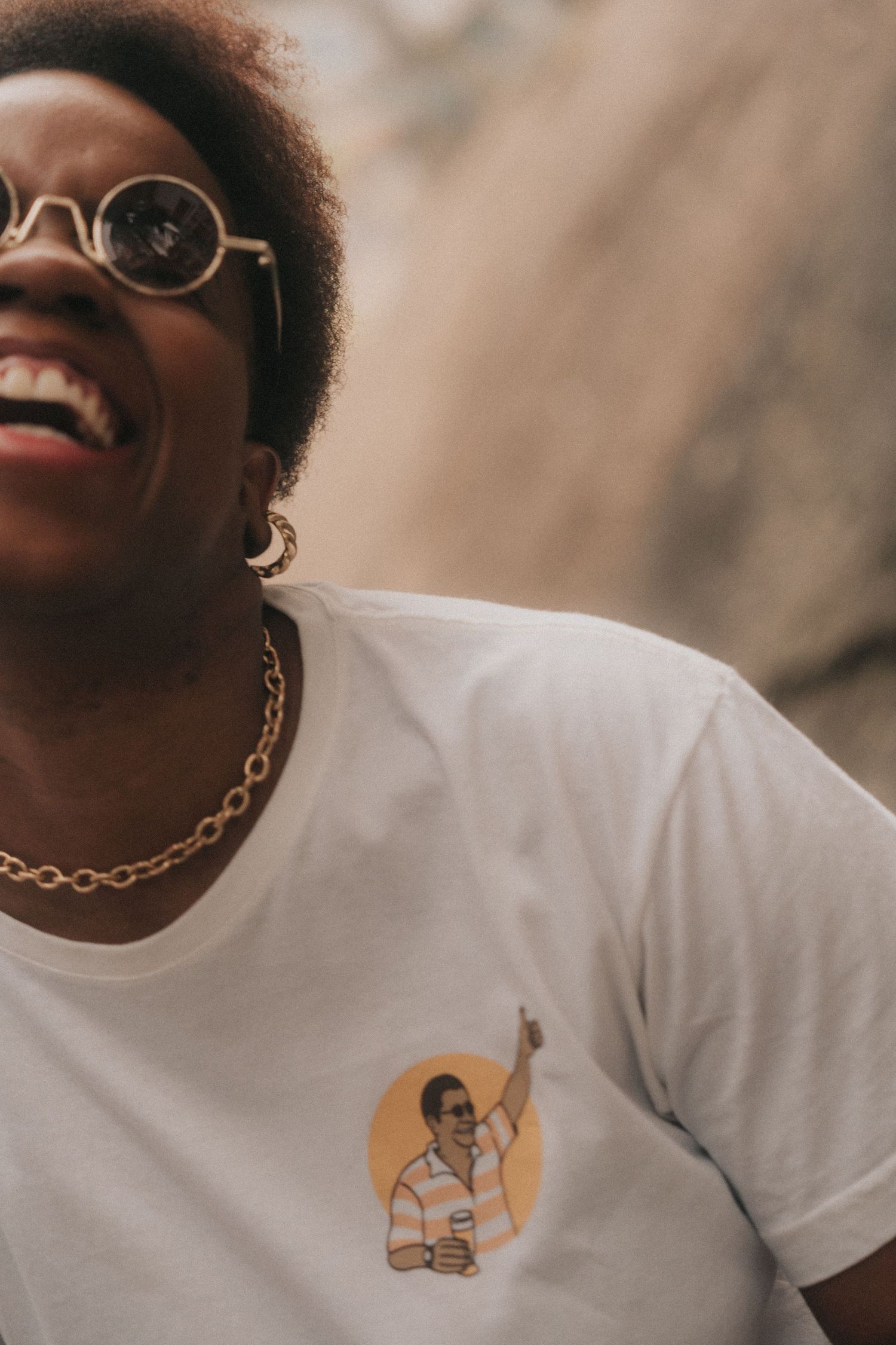 Someone wearing a white t-shirt with a small graphic print laughs joyfully while wearing sunglasses and a gold chain necklace.