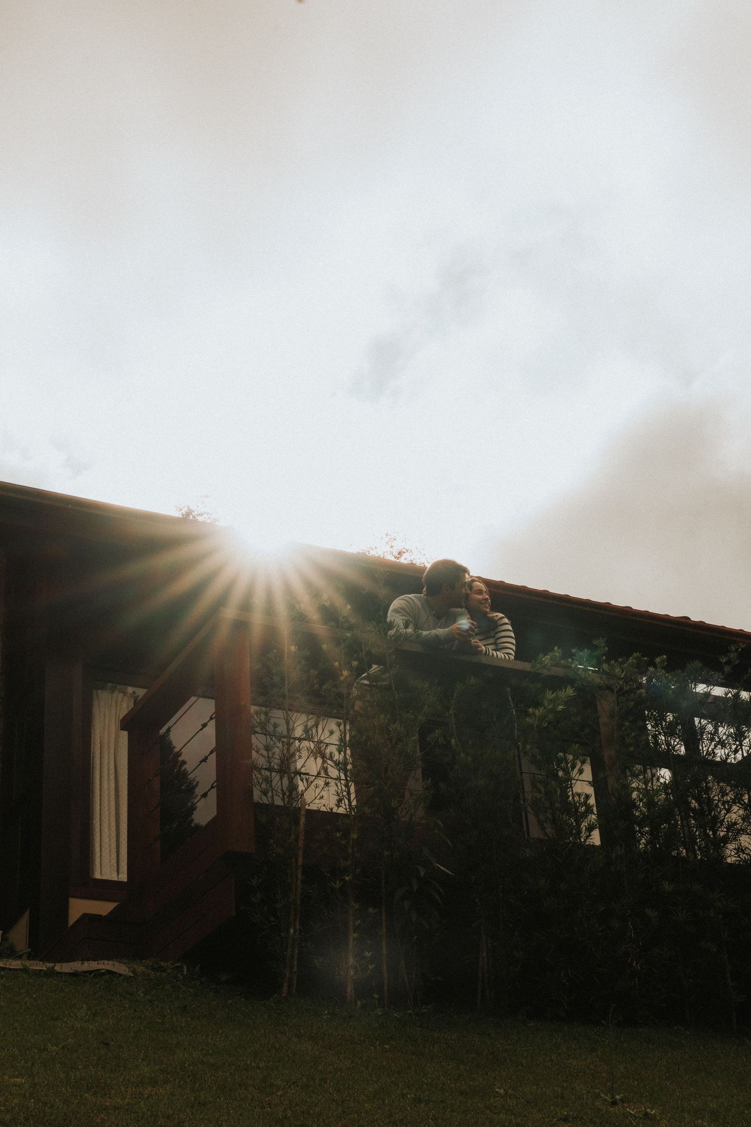Sun rays pierce through dark clouds above silhouetted trees and a house in a moody landscape composition.