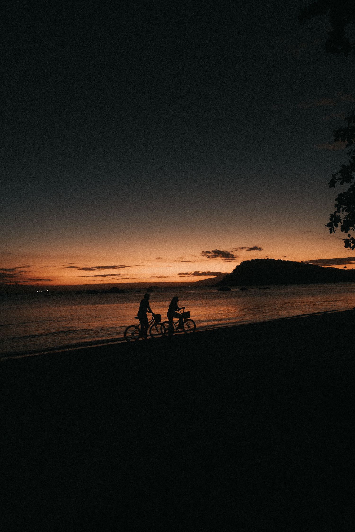 Silhouettes of two cyclists riding along a beach at sunset with mountains in the background.