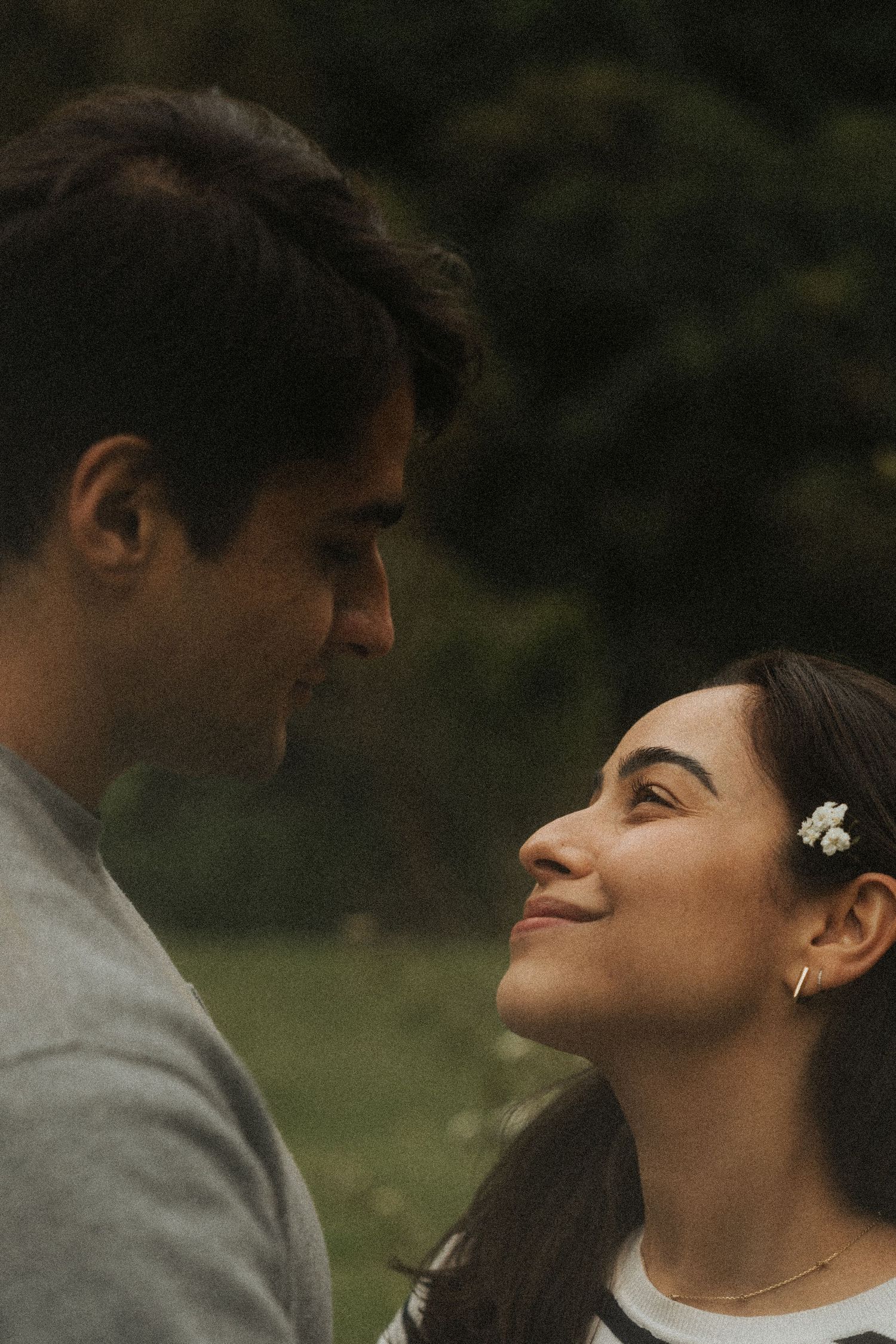 A romantic couple shares an intimate moment outdoors with a small white flower in her dark hair.