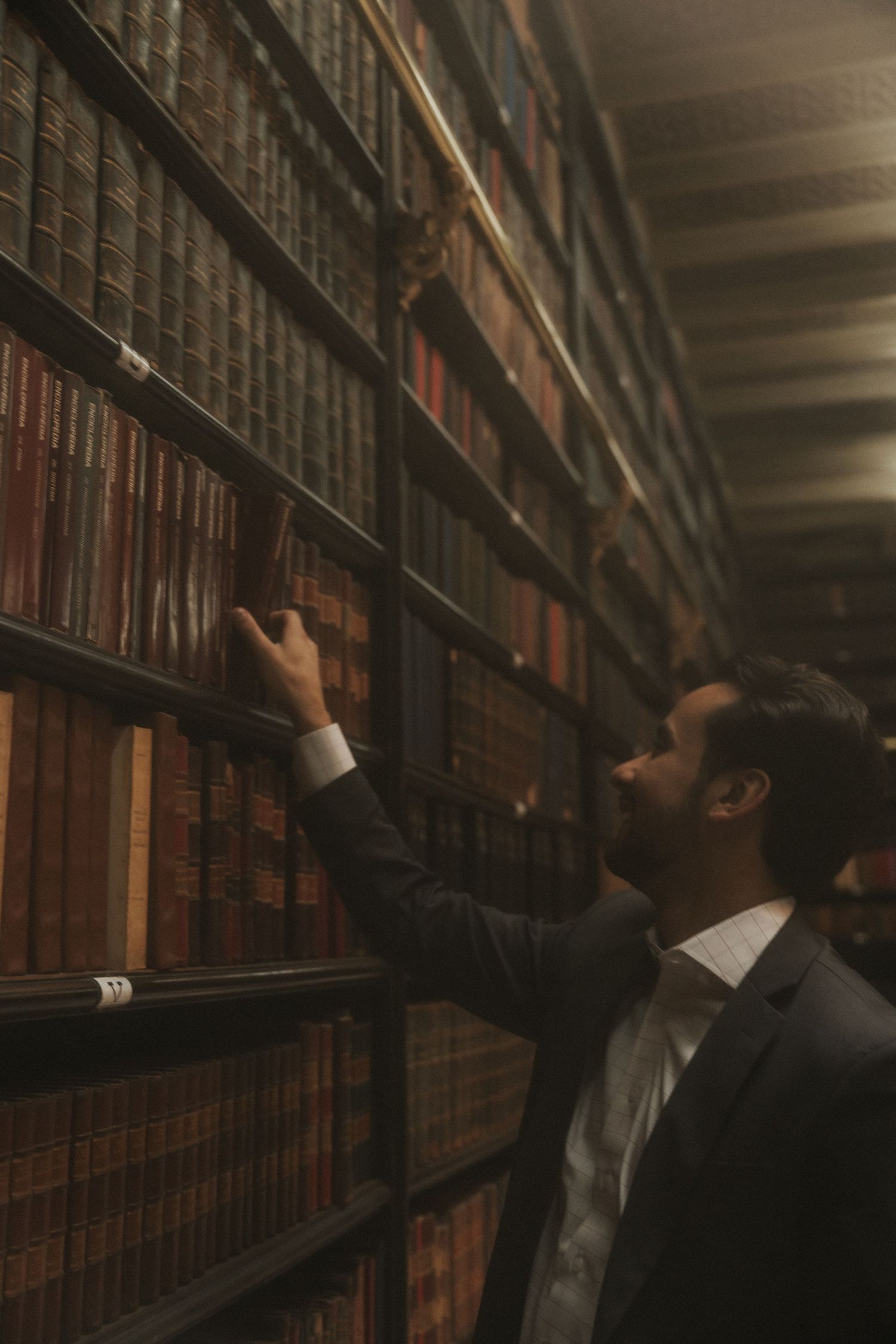Someone reaches for an old book in a dimly lit library with towering wooden shelves filled with vintage volumes.