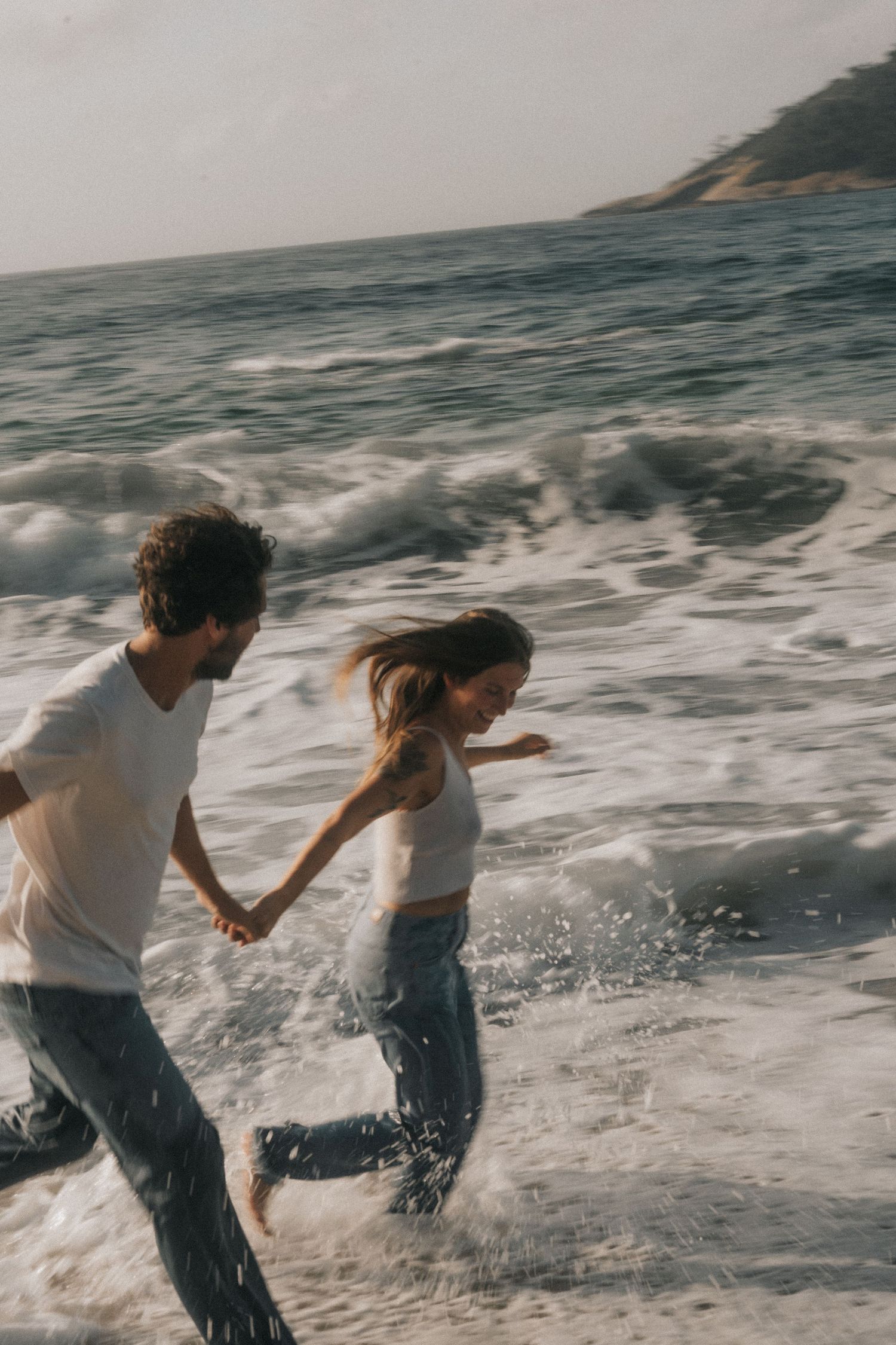 Two people run through ocean waves along the beach while splashing in the water on a sunny day.
