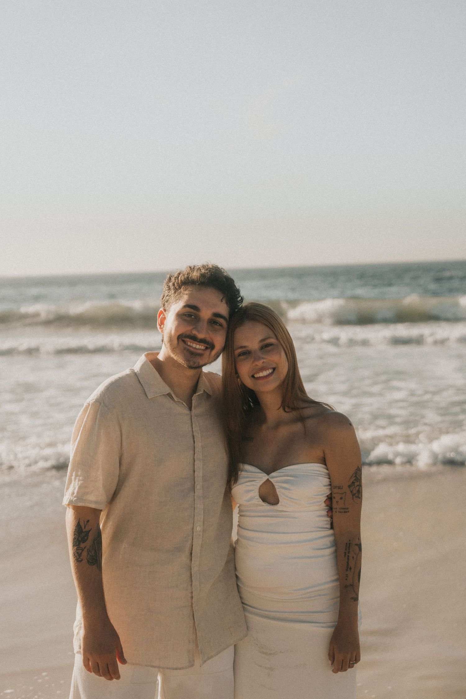 A couple enjoys a romantic moment together at the beach during sunset in matching beige and white outfits.