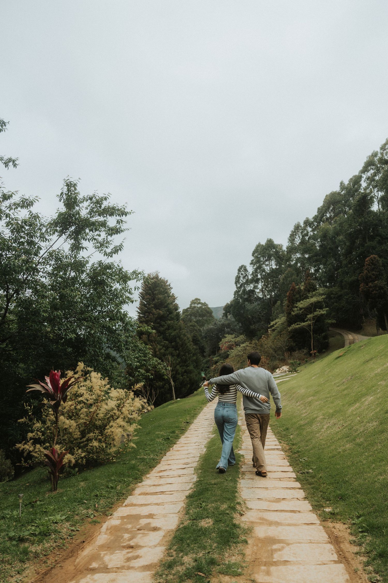 A person walks along a winding stone path through a green hillside garden on an overcast day.