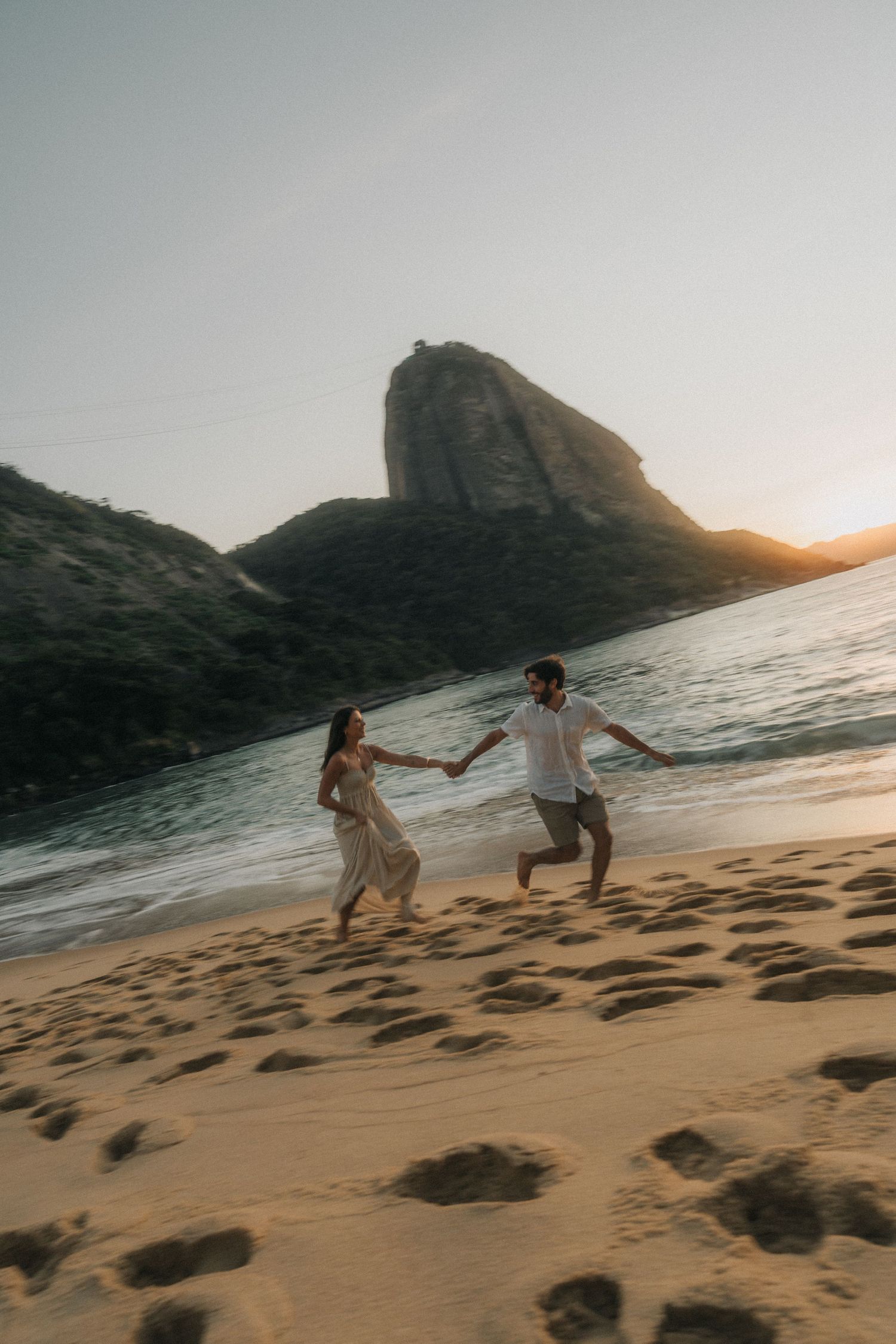 Couple running playfully along a scenic beach at sunset with rocky cliffs in the background.