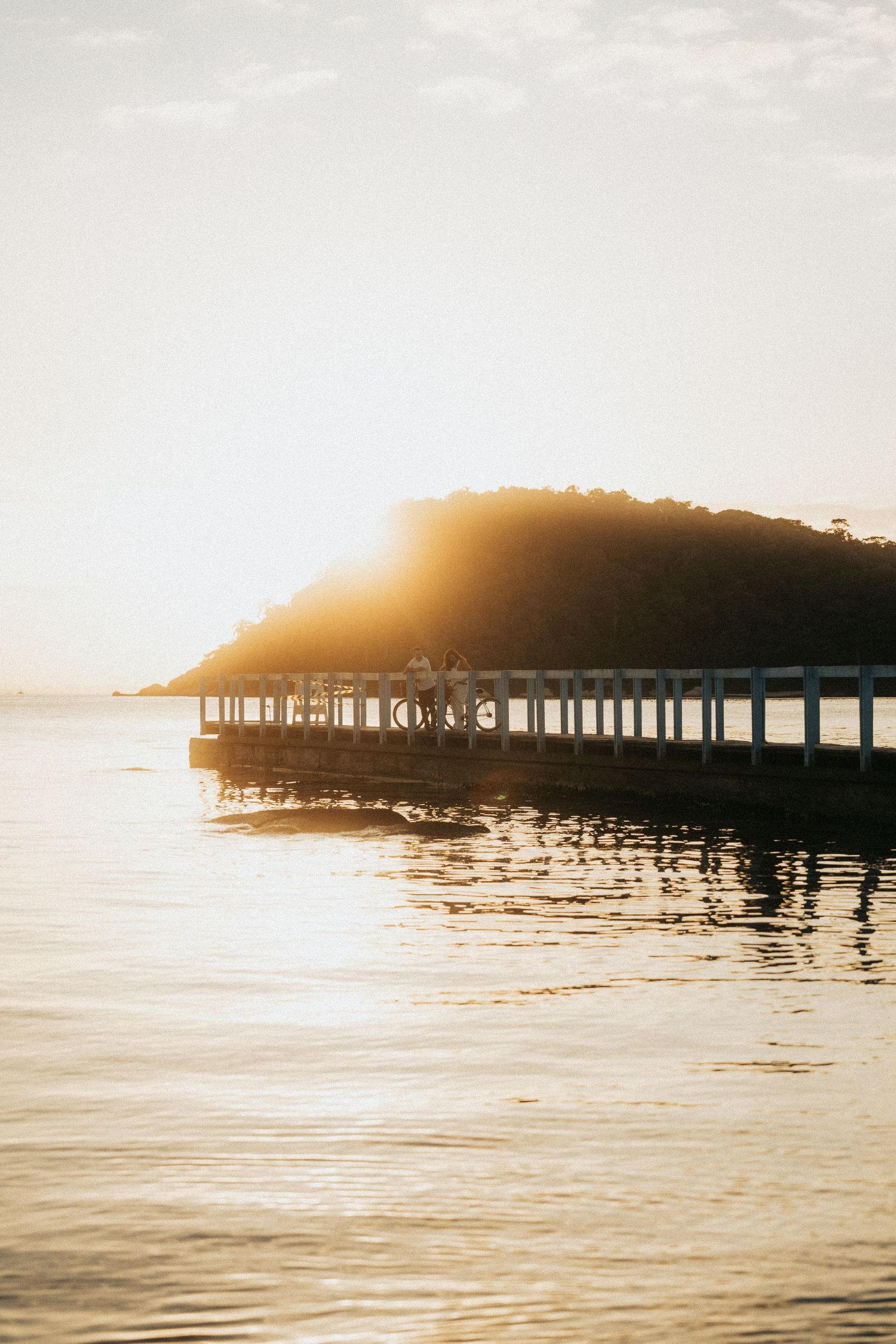 Beautiful sun flares illuminate a wooden pier extending over calm ocean waters at sunset.