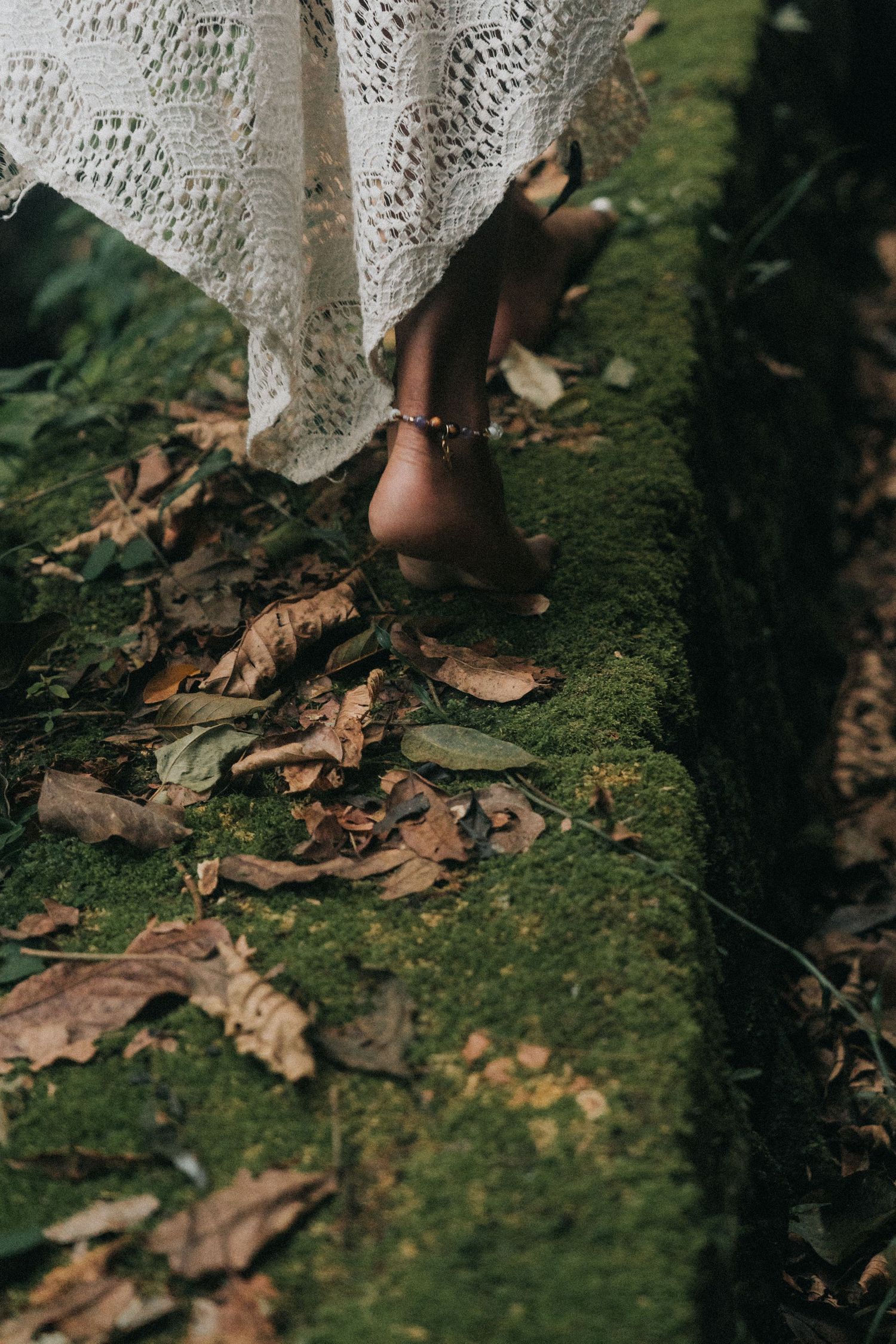 Close-up view of bare feet in lace dress walking on mossy ground with scattered autumn leaves.