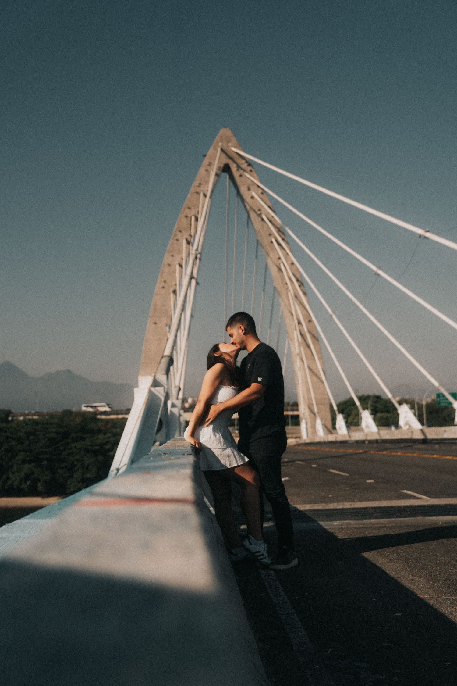 Romantic couple shares intimate moments against modern bridge architecture during sunset photoshoot.