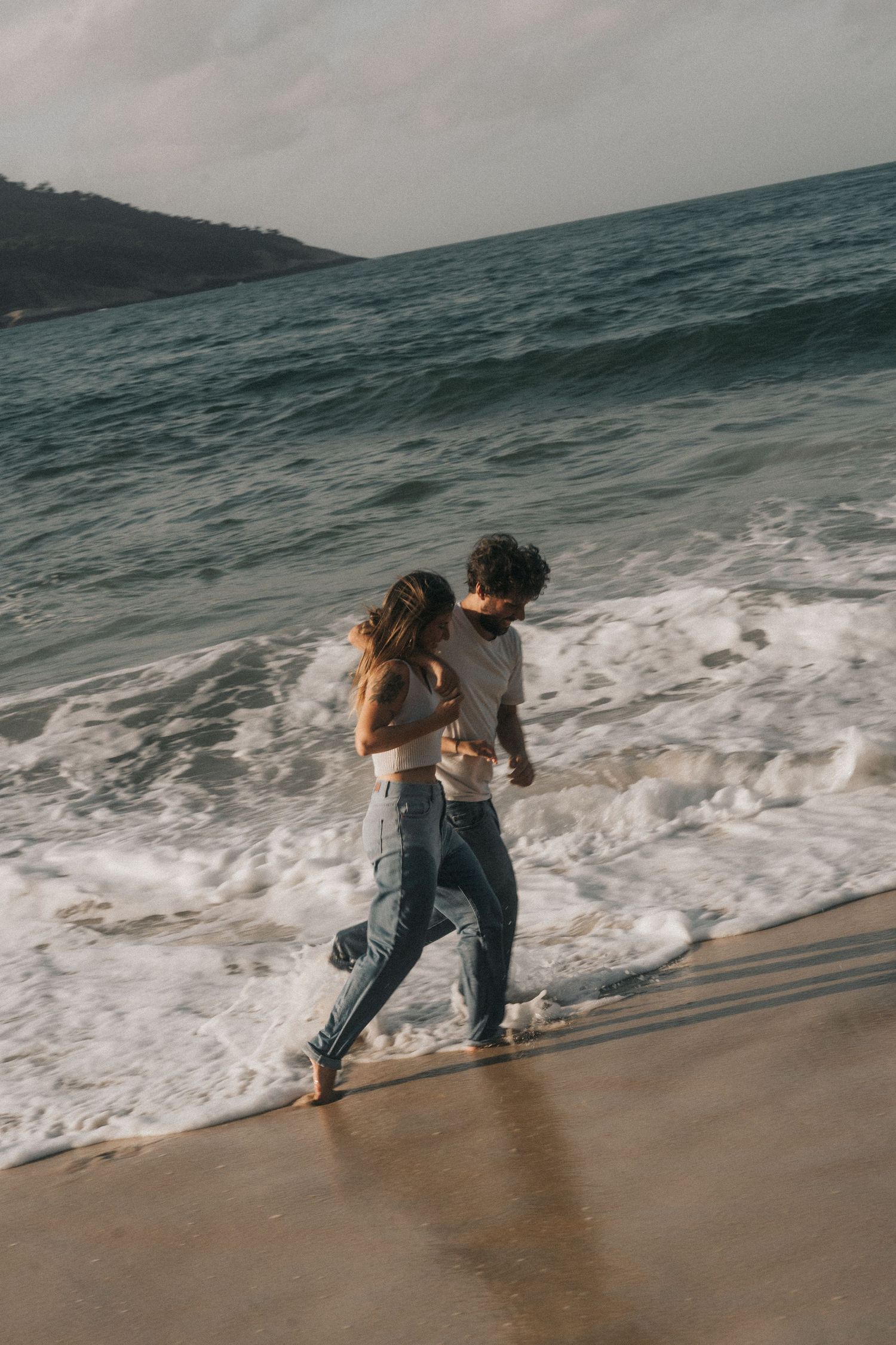 A couple runs along the shoreline as ocean waves crash on a moody beach at sunset.