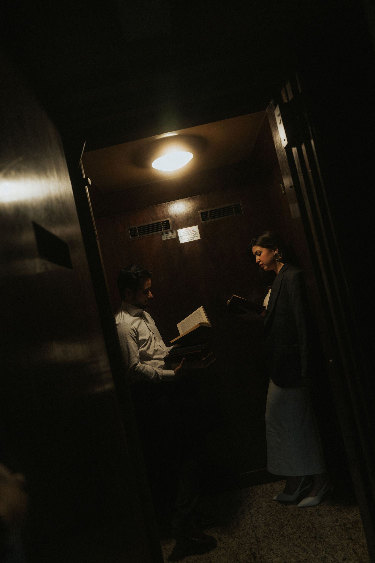 Two people stand in a dimly lit hallway reading documents under a ceiling light in moody vintage atmosphere.