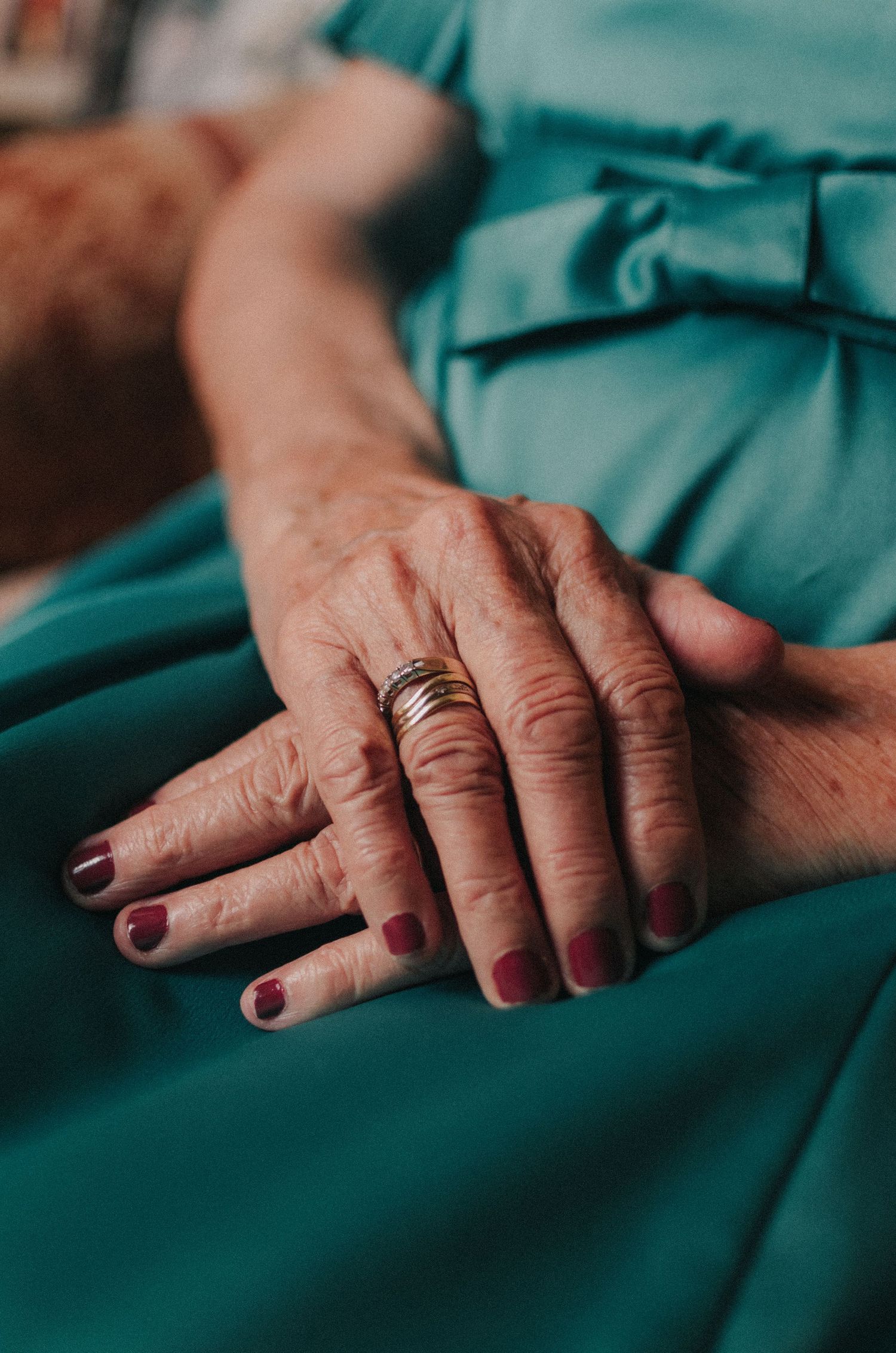 Close-up view of weathered hands with painted nails resting on teal fabric, showing multiple rings on fingers.