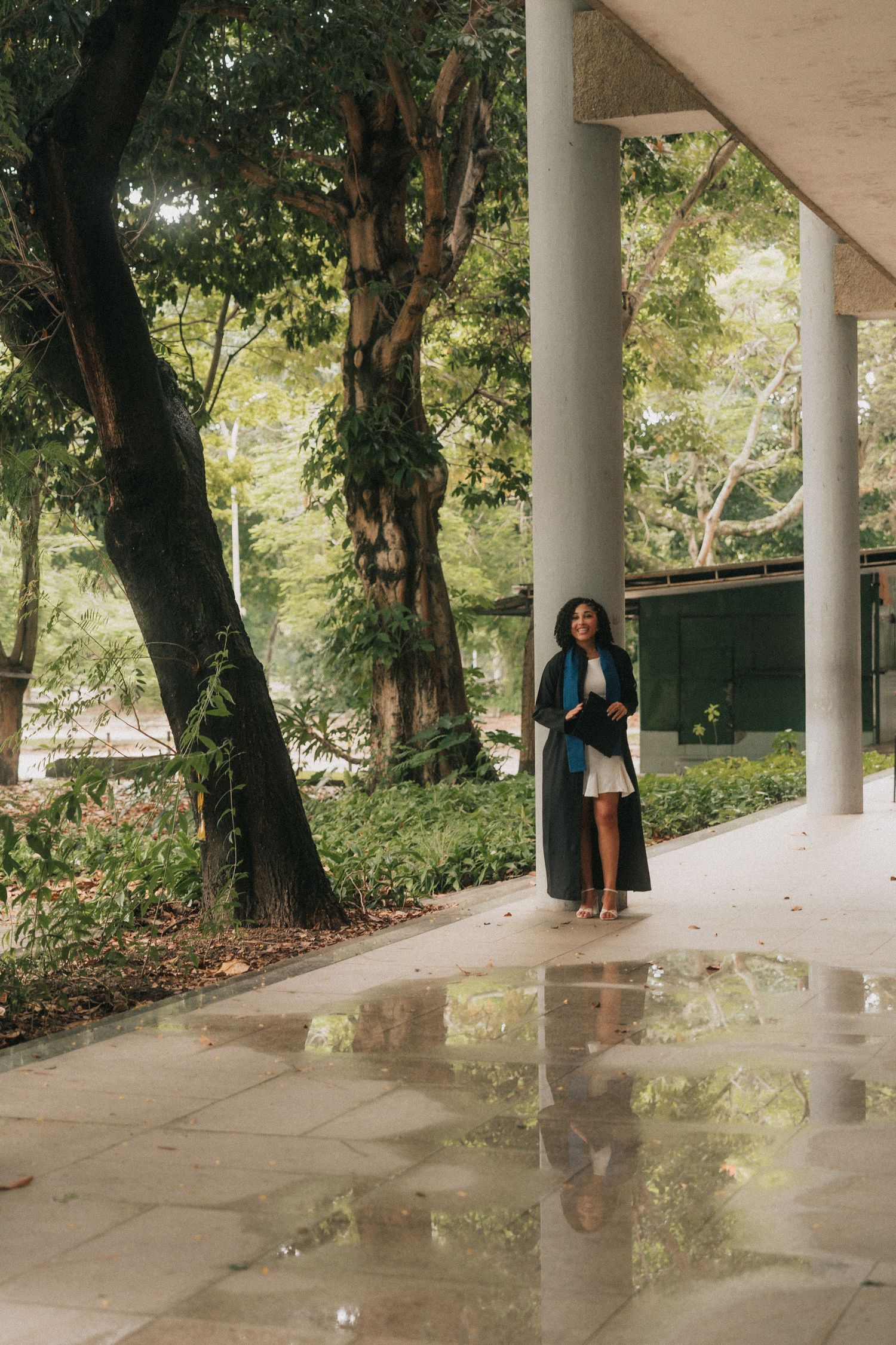 People walking under a covered walkway with stone pillars and puddles reflecting greenery on a rainy day.