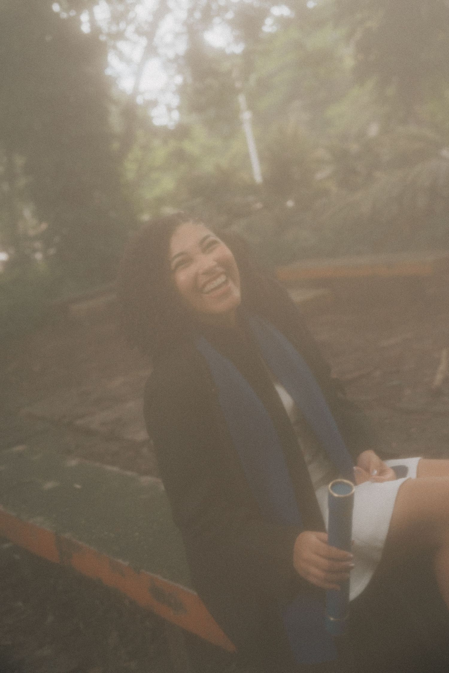 A person in a navy blazer sits outdoors holding a notebook and laughing joyfully on a misty day.