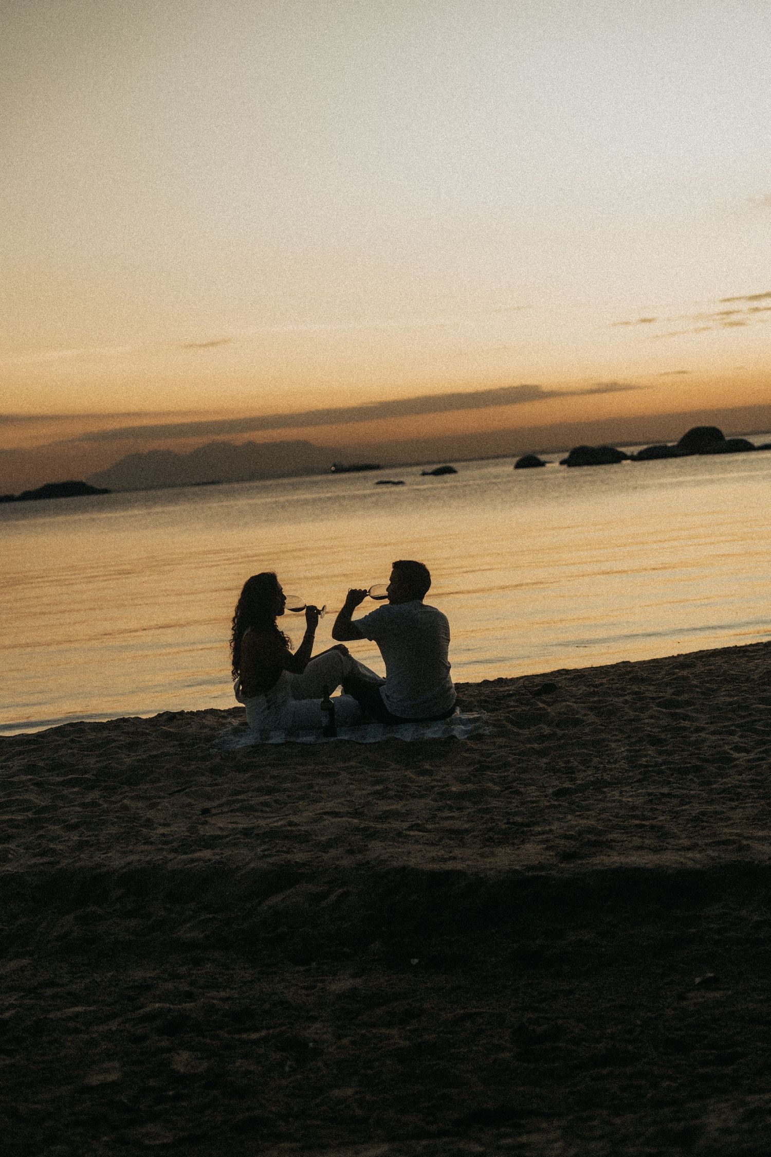 Silhouettes of people sitting on a beach at sunset with mountains in the distance and golden sky reflections on the water.